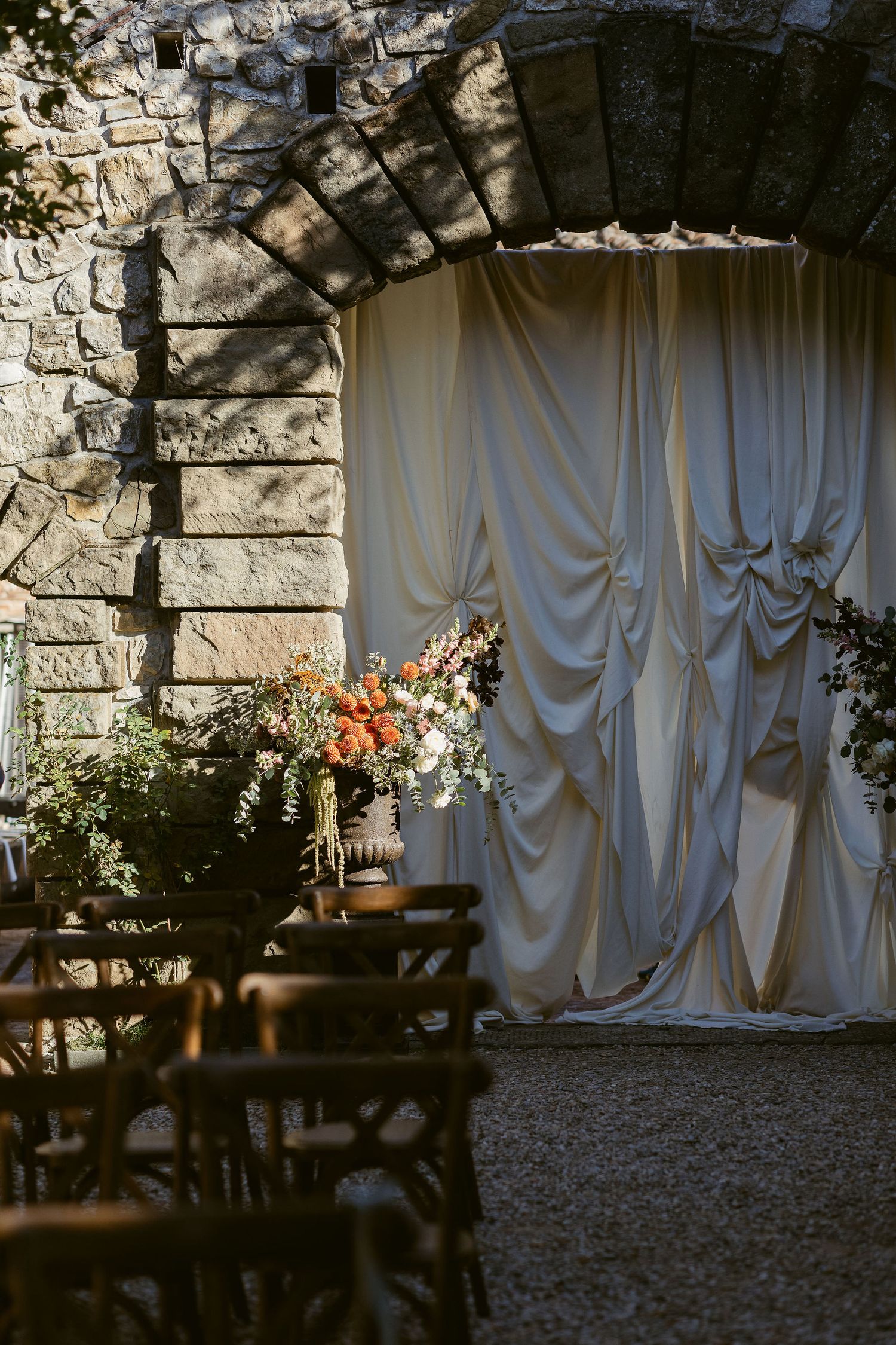 Stone archway with draped white curtains and floral decorations creating an elegant outdoor event setting.