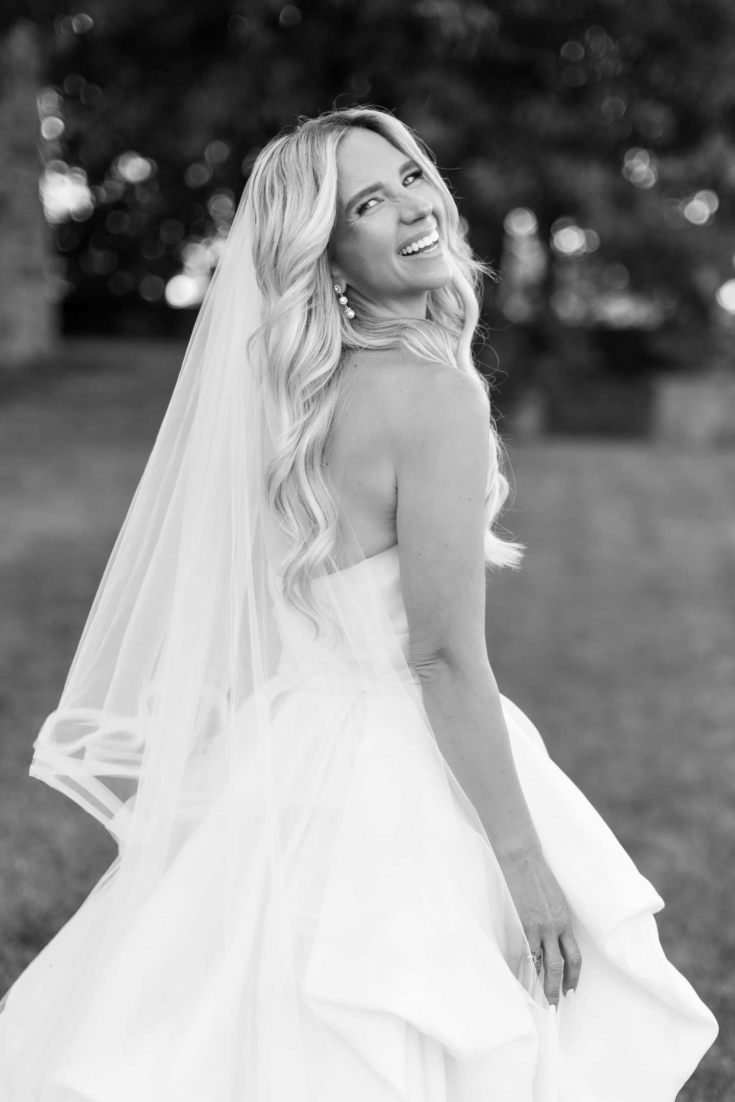 A joyful bride in a flowing white wedding dress and veil poses outdoors with a natural candid expression.