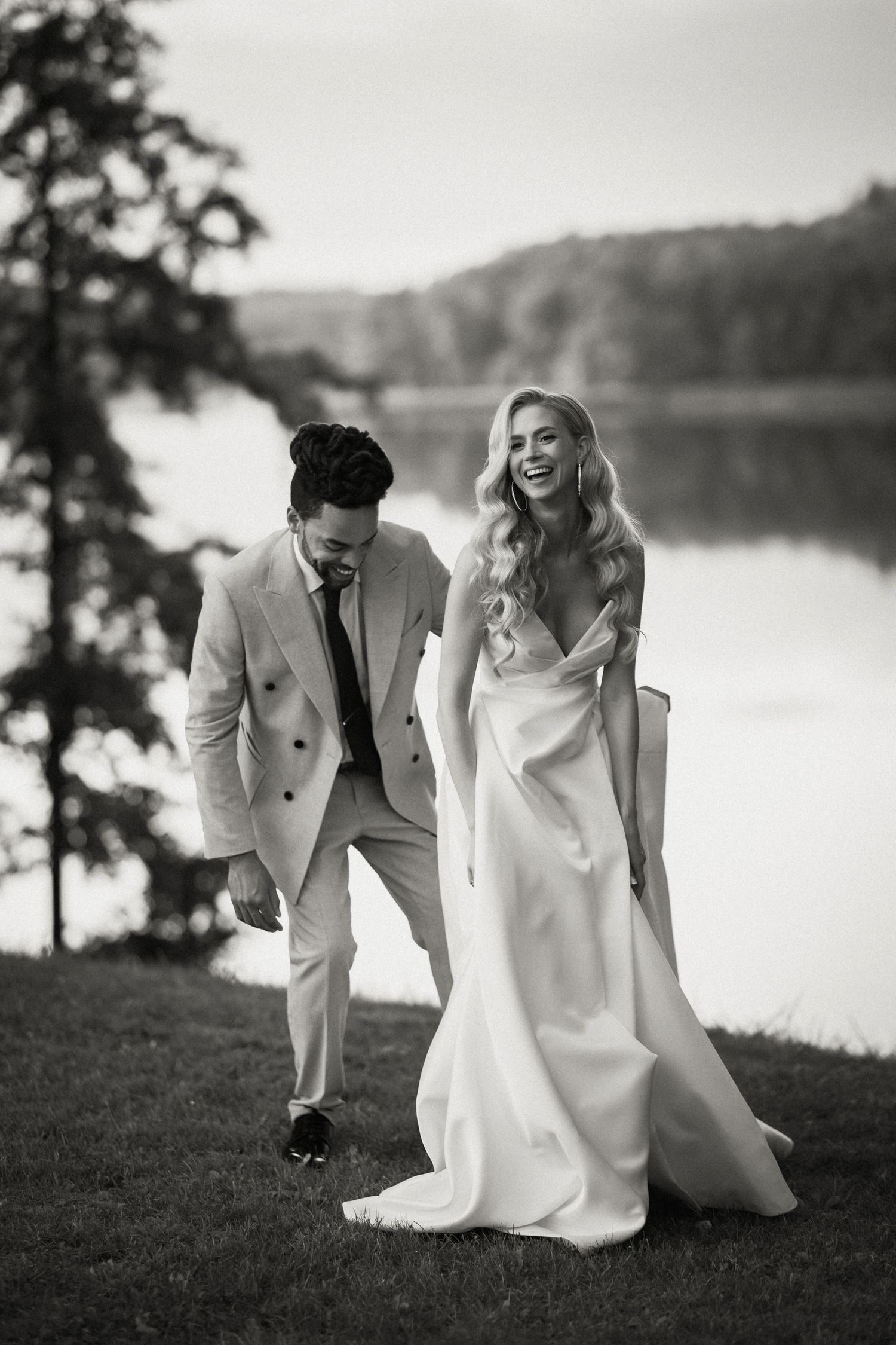 A joyful candid wedding photo in black and white showing a couple laughing by a serene lakeside.