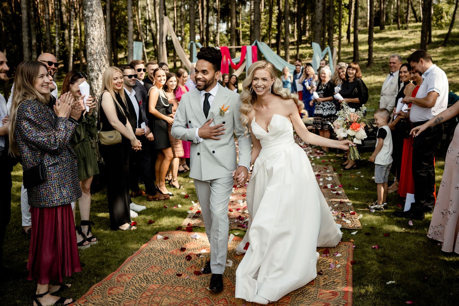 Wedding recessional along rustic carpet runner in forest with guests celebrating.
