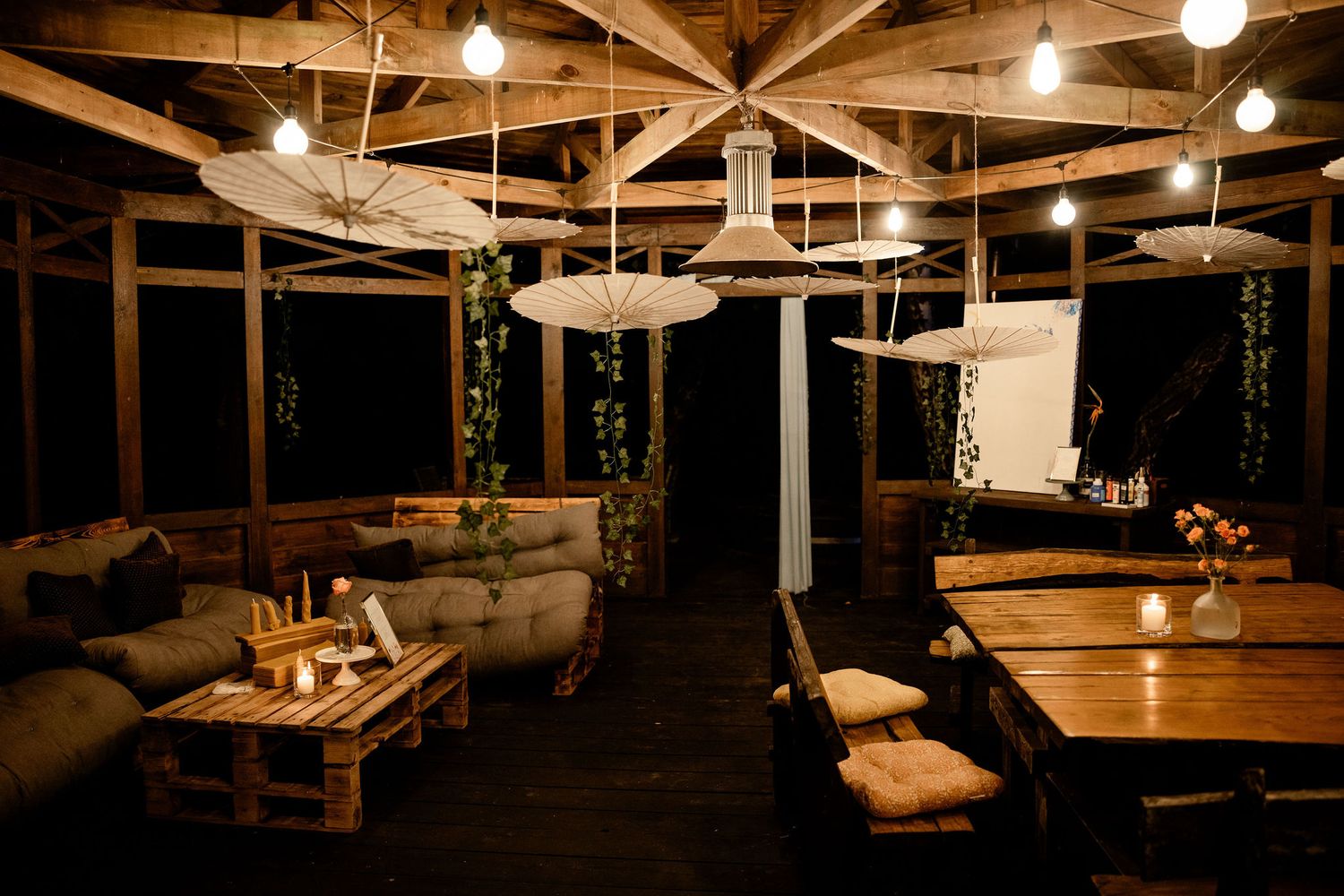 Cozy rustic indoor-outdoor patio space with warm lighting, lounge furniture, and wooden beams at night.