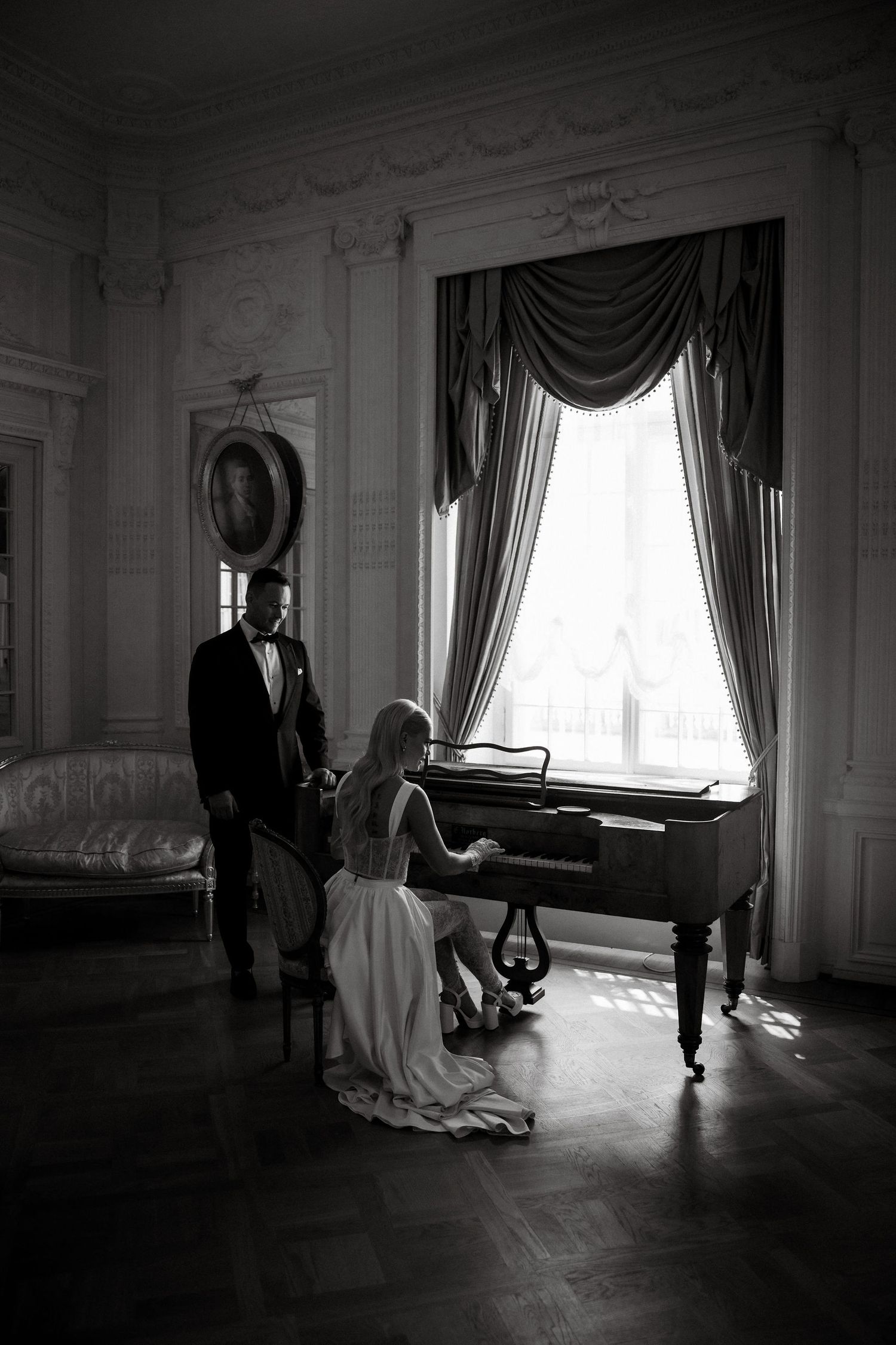 Black and white photo of a couple at a grand piano in an elegant room with dramatic curtains.