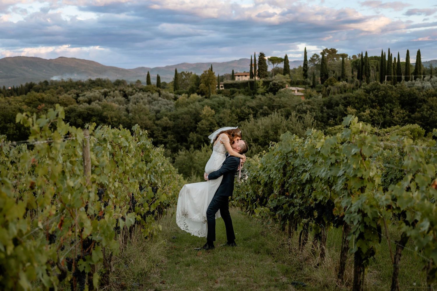 Romantic couple embrace among vineyard rows with cypress trees and mountains in the background.