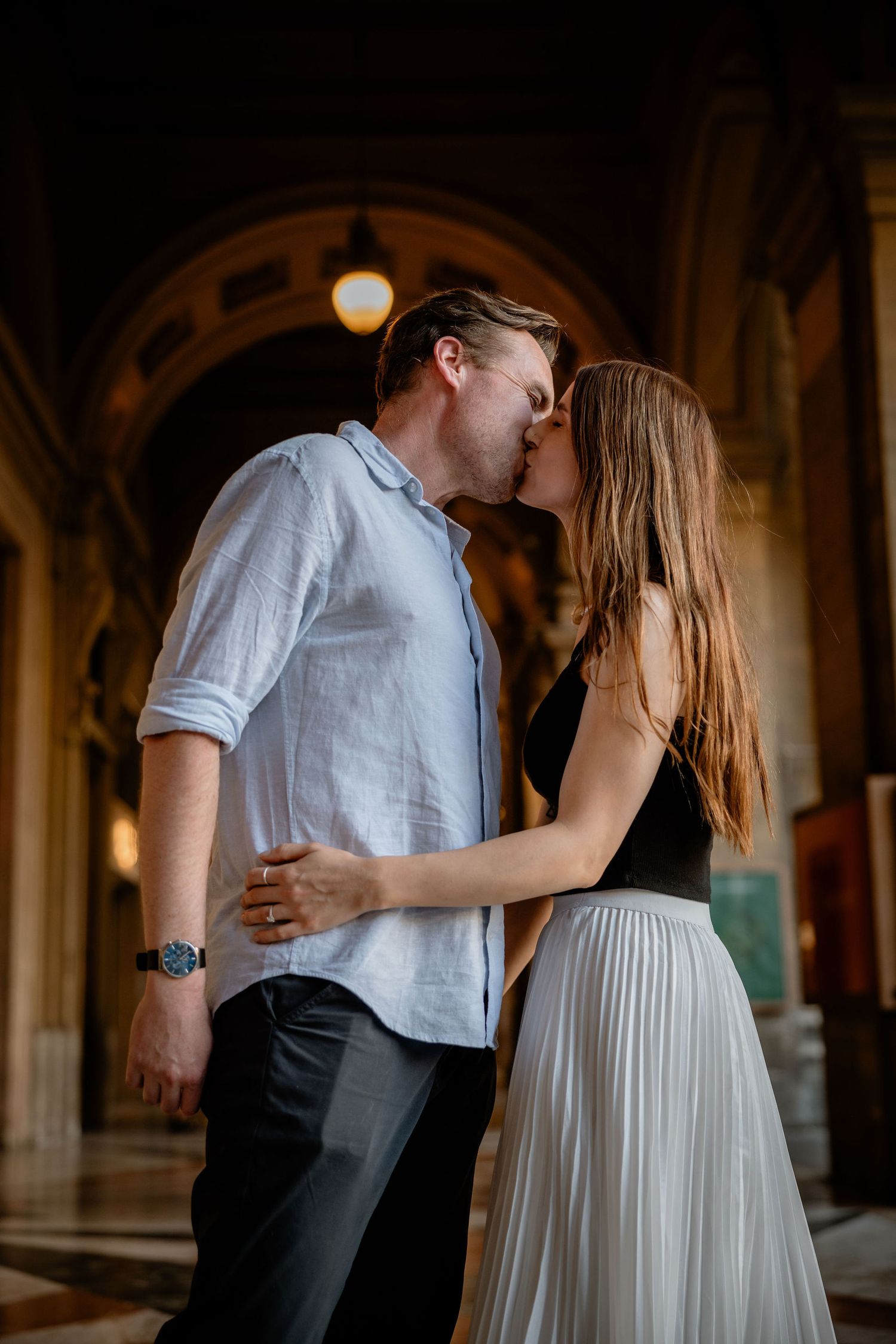A romantic couple shares a tender kiss under an arched passageway lit by a warm glowing light fixture.