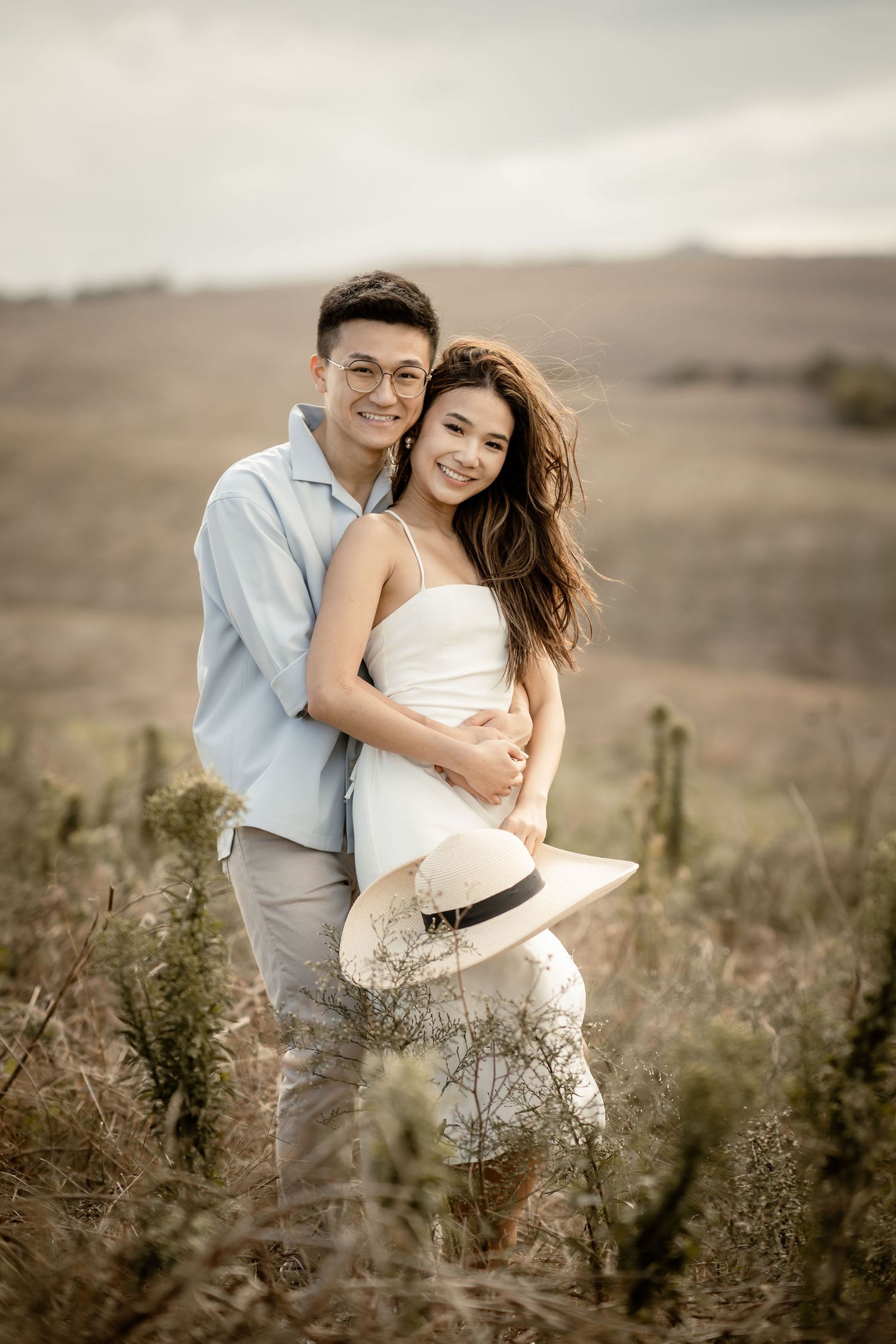 A couple in coordinated outfits embraces while holding a straw hat in a rustic landscape.