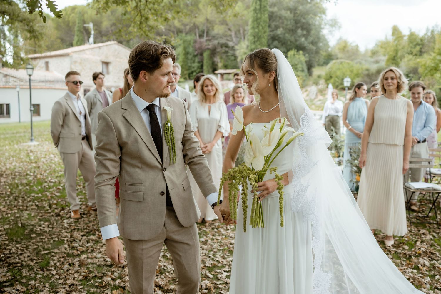 Wedding party in neutral tones celebrates and laughs together outdoors under trees during a joyful moment.