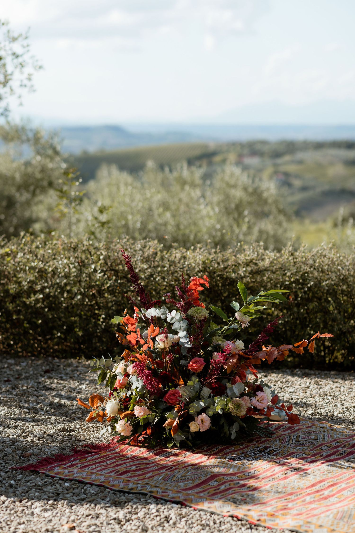 A colorful floral arrangement sits on a rustic blanket against a scenic hillside backdrop on a sunny day.
