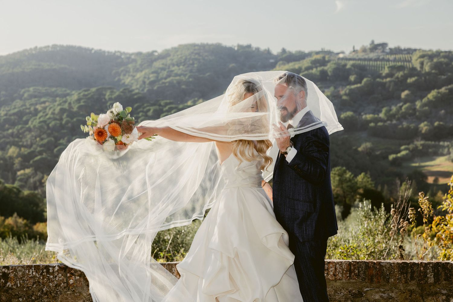 A flowing white wedding dress veil catches the wind against a scenic mountain landscape backdrop at sunset.