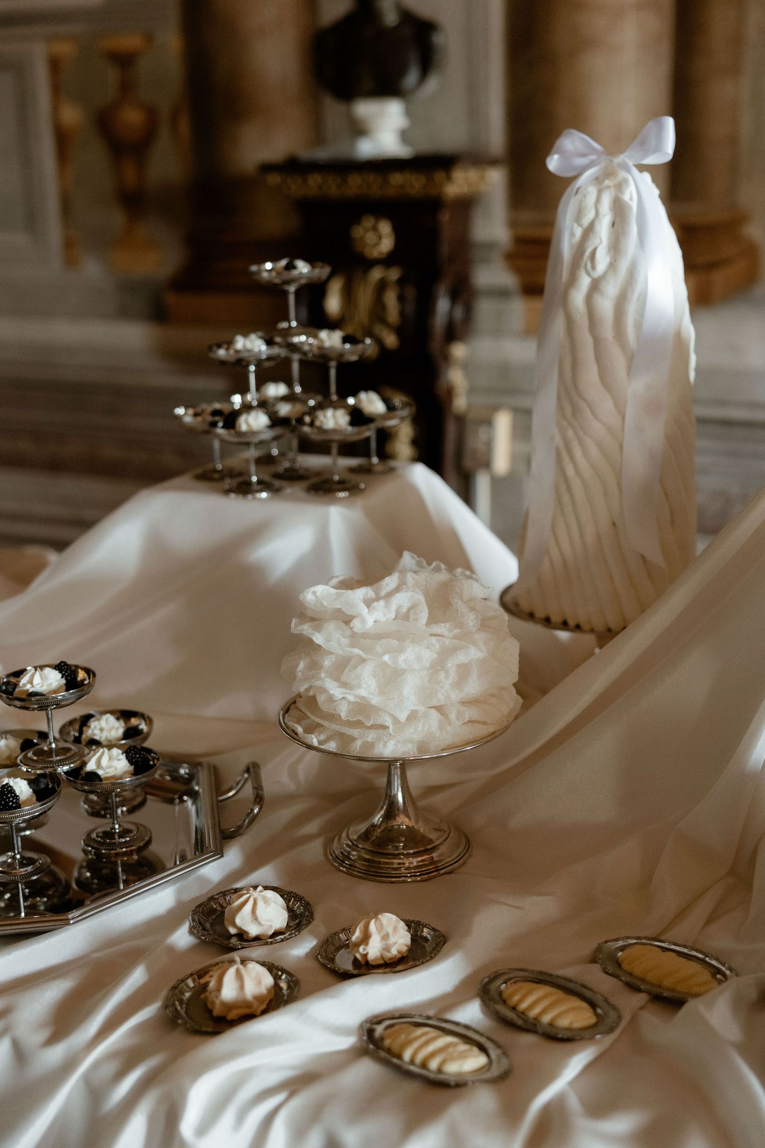 Elegant white wedding dessert display with layered cake and cupcakes on silver stands atop silky white tablecloth.