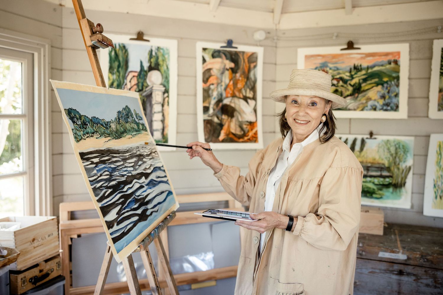 Artist in beige coat works on abstract landscape painting while standing at wooden easel in bright studio with artwork on walls.