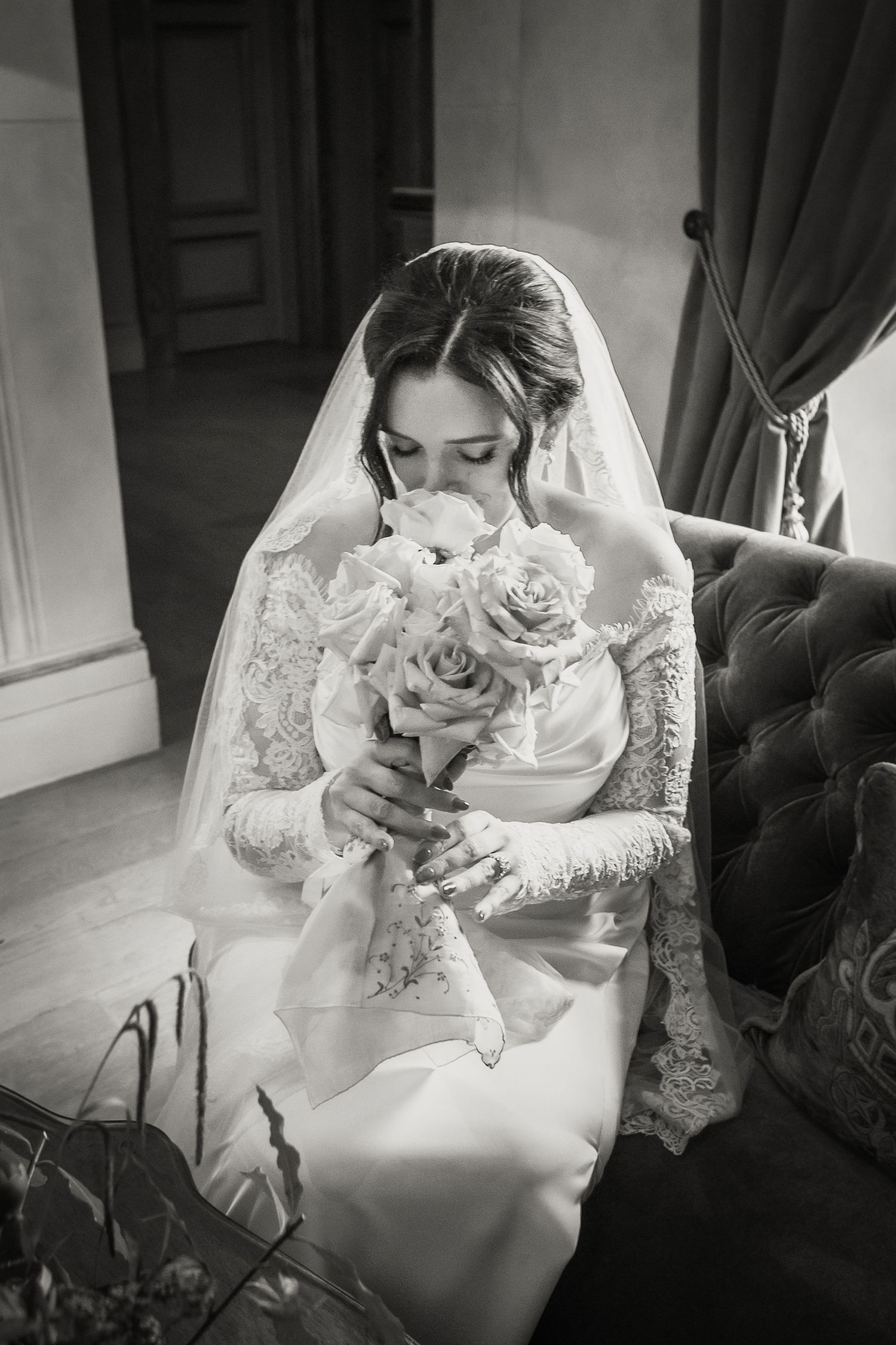 Vintage black and white photo of bride in lace gown smelling elegant rose bouquet while seated on velvet chair.