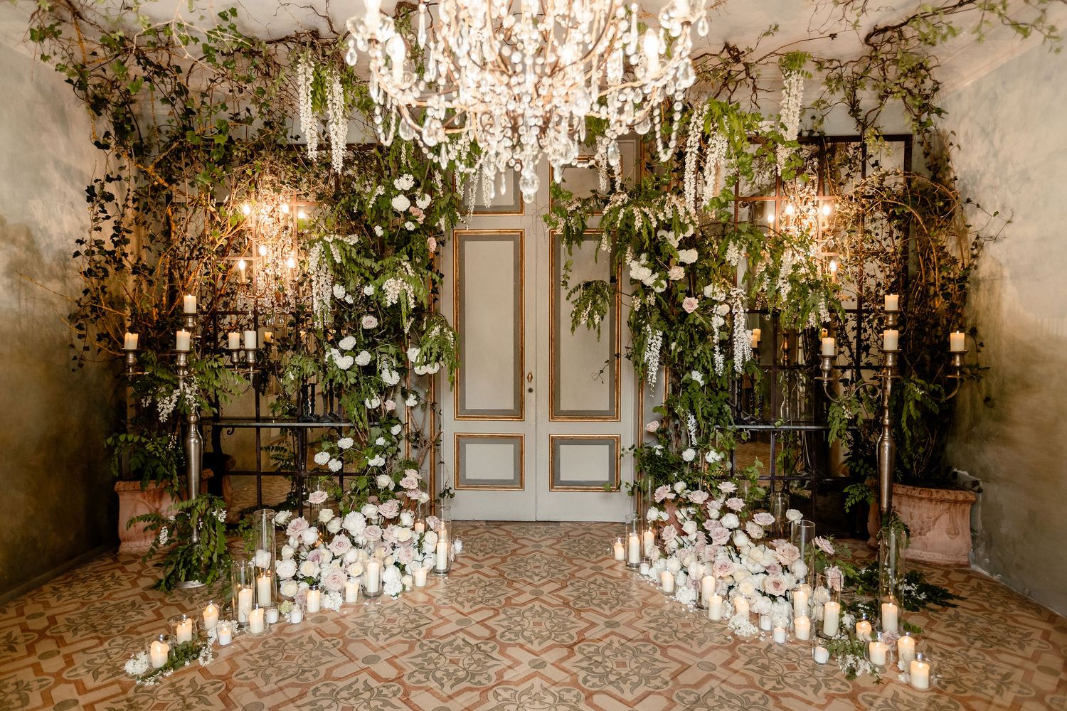 Elegant wedding venue entrance with crystal chandeliers and cascading greenery surrounded by glowing candles on a stone floor.