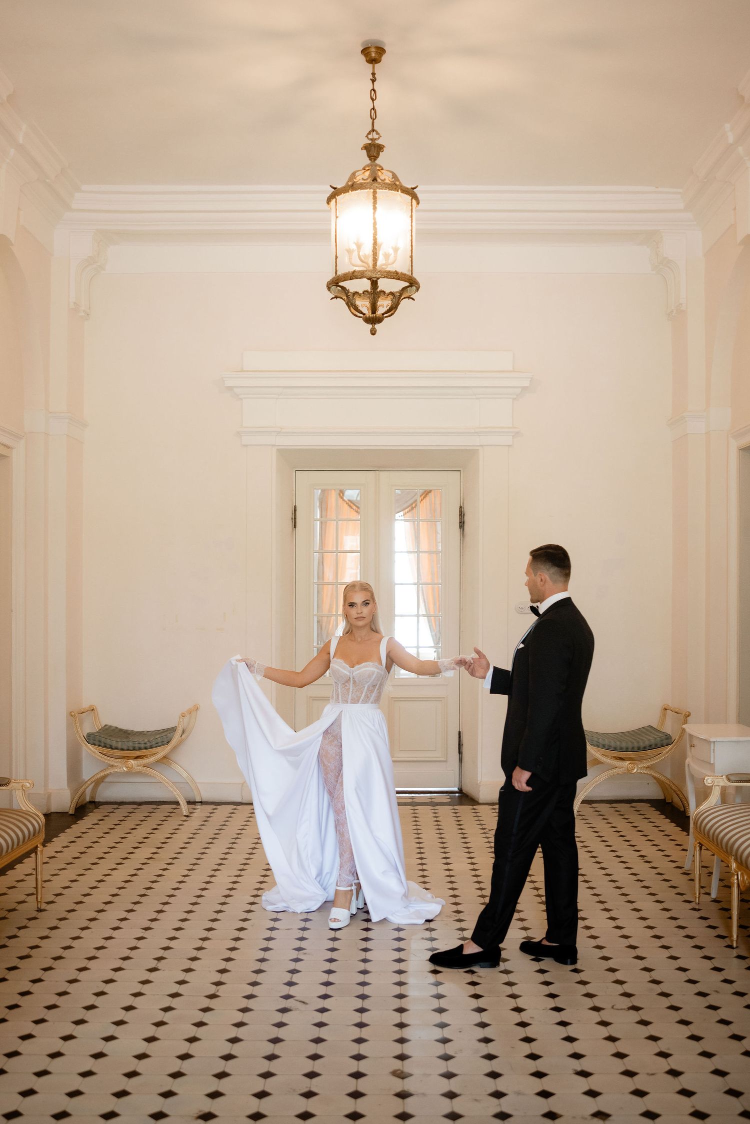 A couple poses elegantly in a grand foyer with a vintage chandelier and checkered floor.
