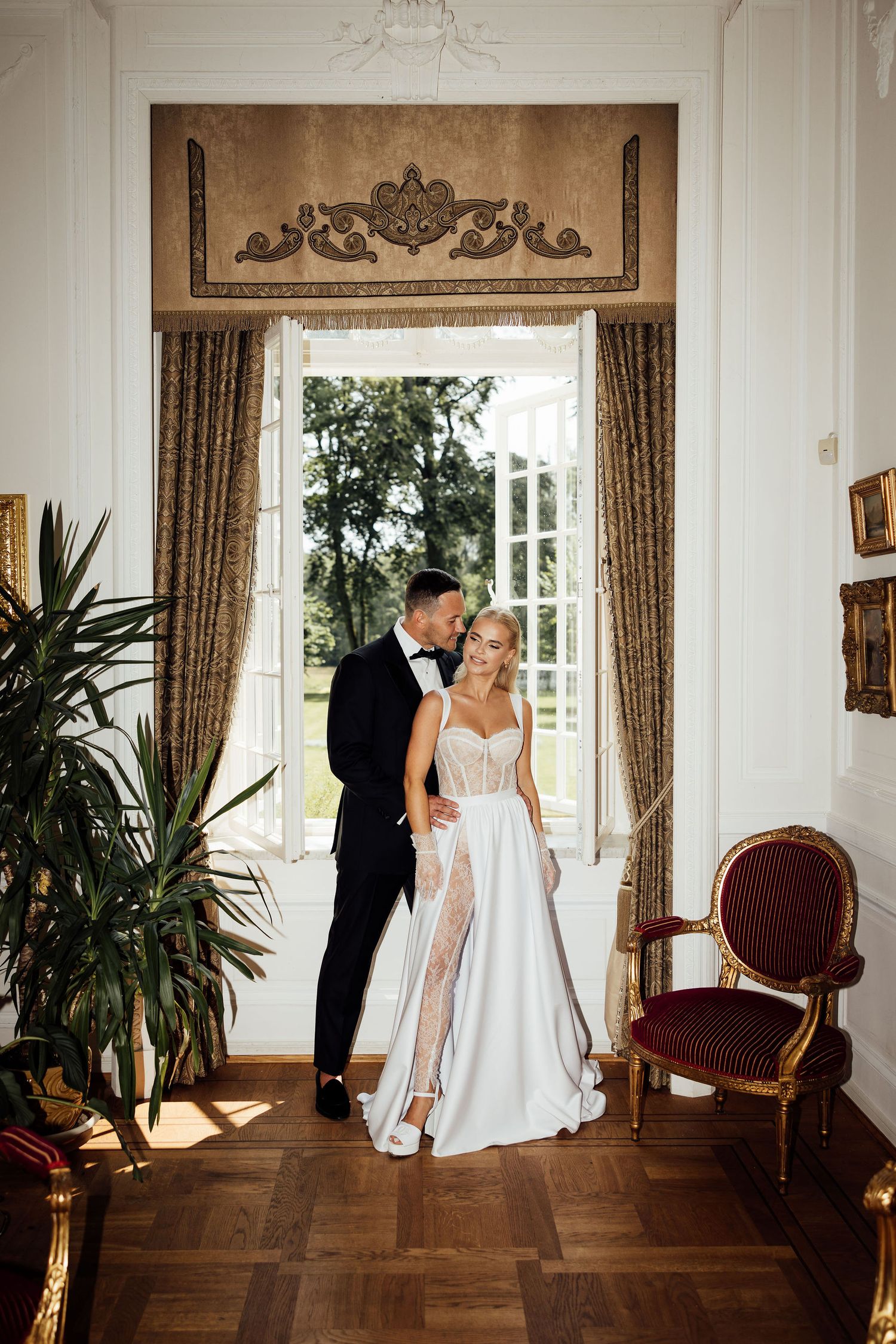 Romantic wedding couple posing in an ornate room with classical decor and French windows.