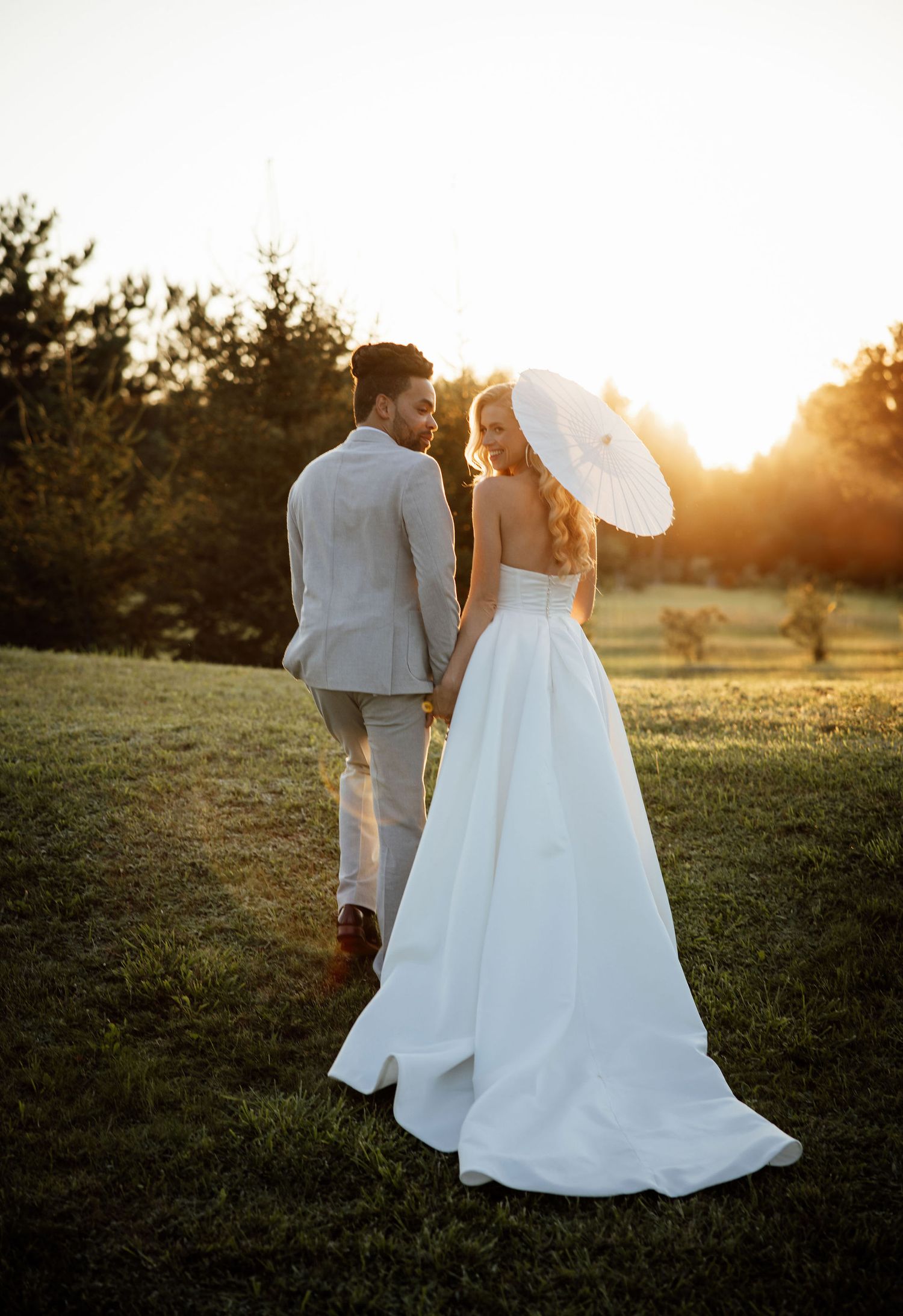 Romantic sunset wedding portrait features a couple with a parasol standing in a golden-lit field against a setting sun.