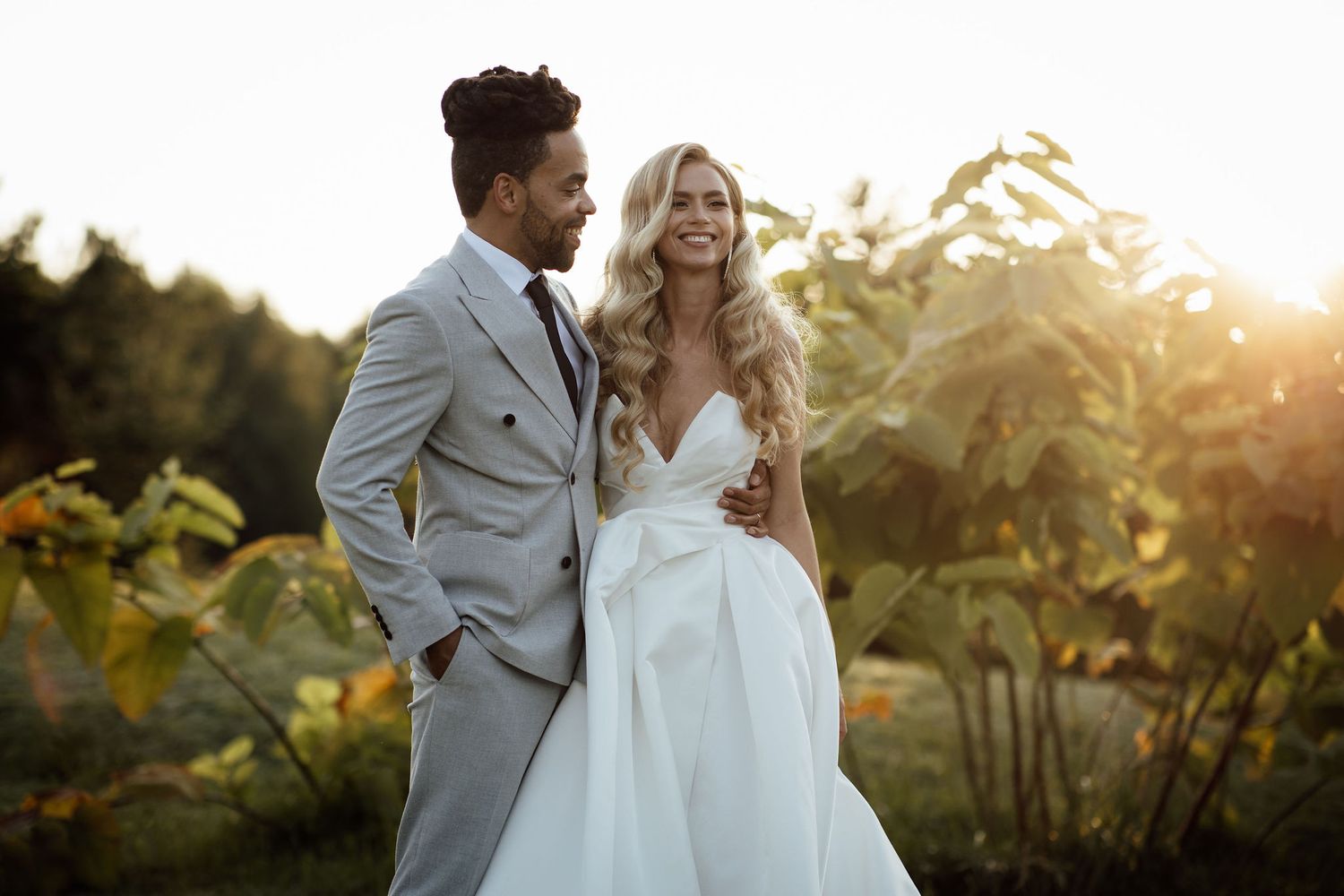 A newlywed couple stands together outdoors at sunset with sunflowers in the background.