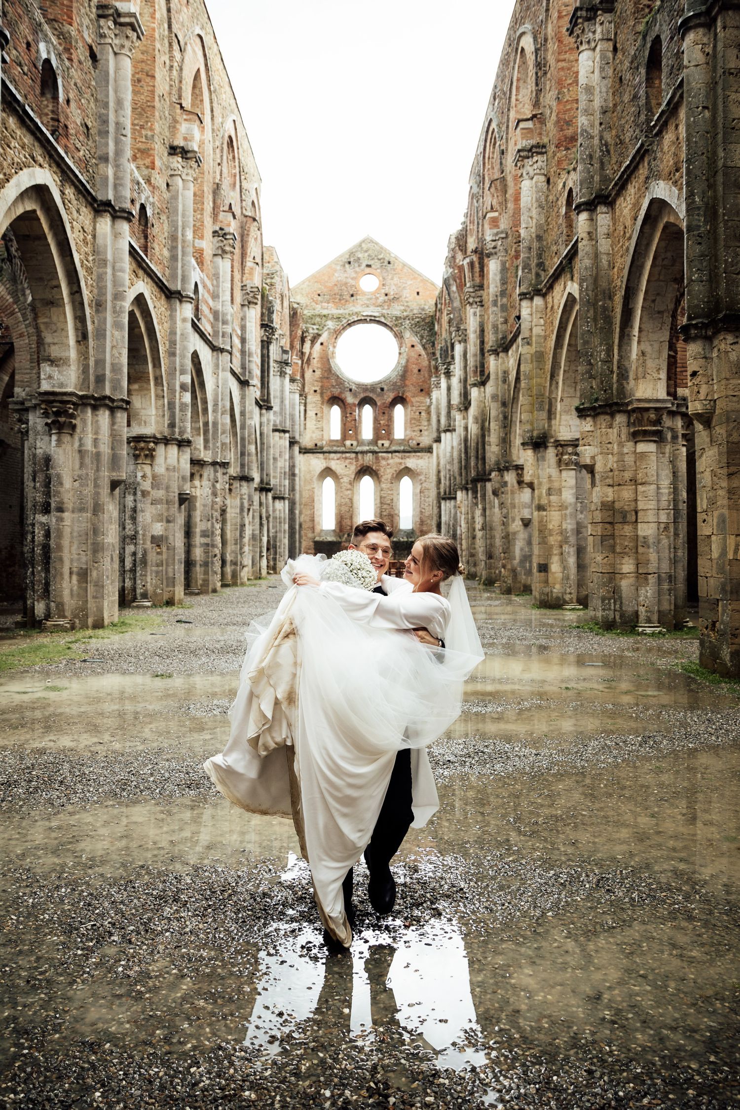 A couple embraces in a romantic moment inside the ruins of a historic stone cathedral with towering Gothic arches. San Galgano.