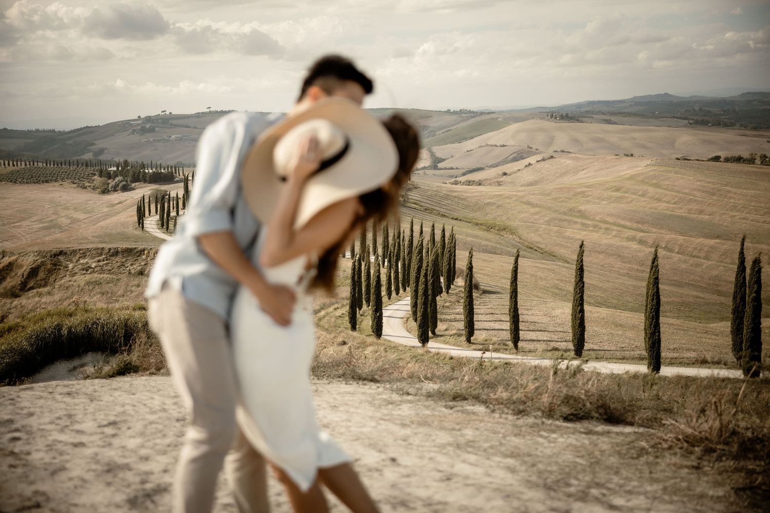 A romantic couple embraces against a backdrop of rolling Tuscan hills lined with cypress trees.
