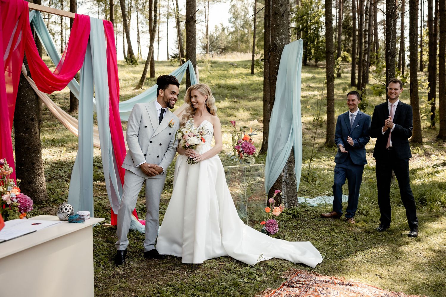 Newlyweds stand under decorative red and blue drapes during their forest wedding ceremony.