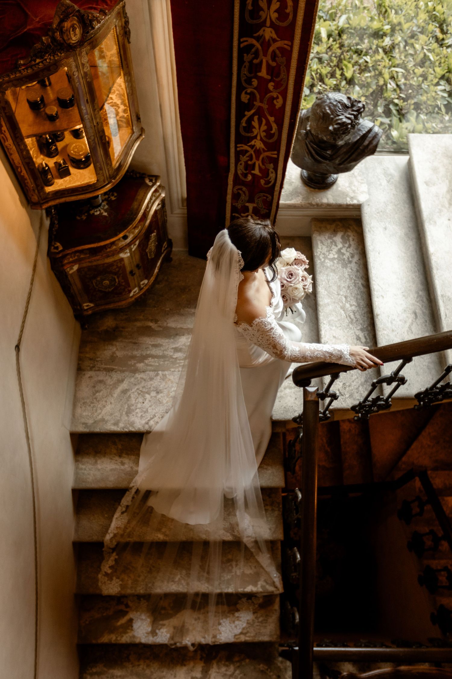 A flowing white wedding dress and veil cascade down vintage stone stairs beside an antique decorative cabinet.