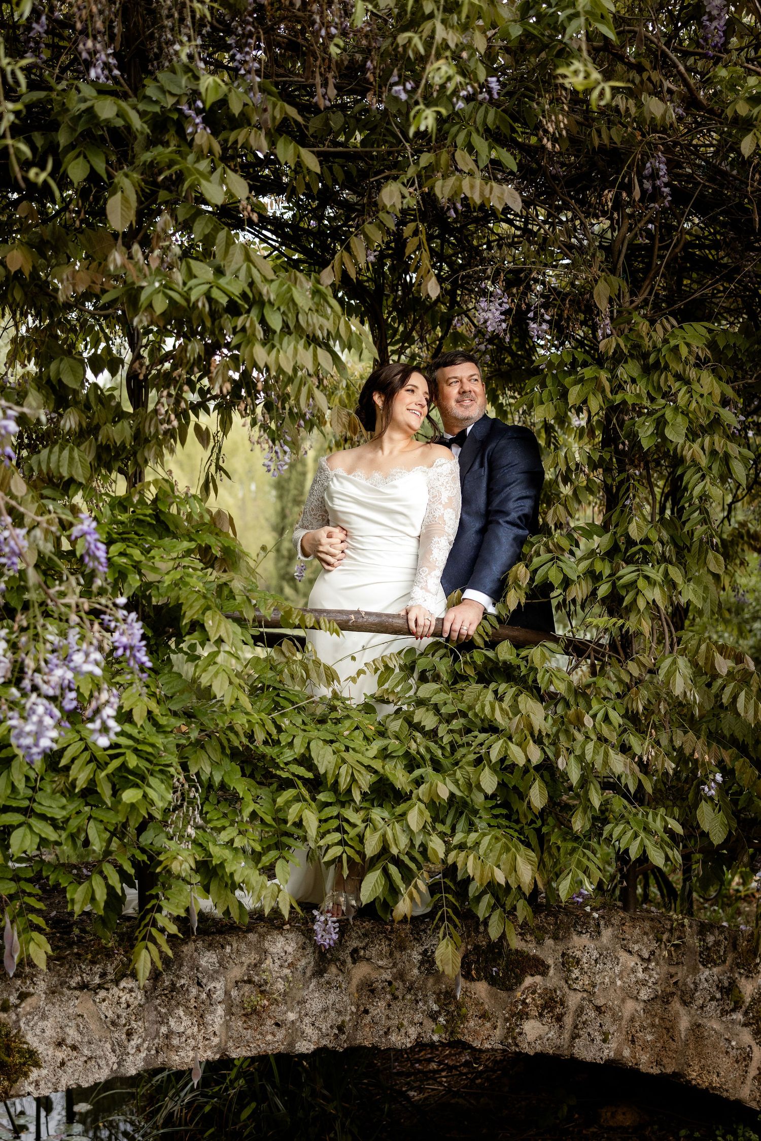 A couple poses together on a rustic stone bridge surrounded by lush greenery and purple flowers.
