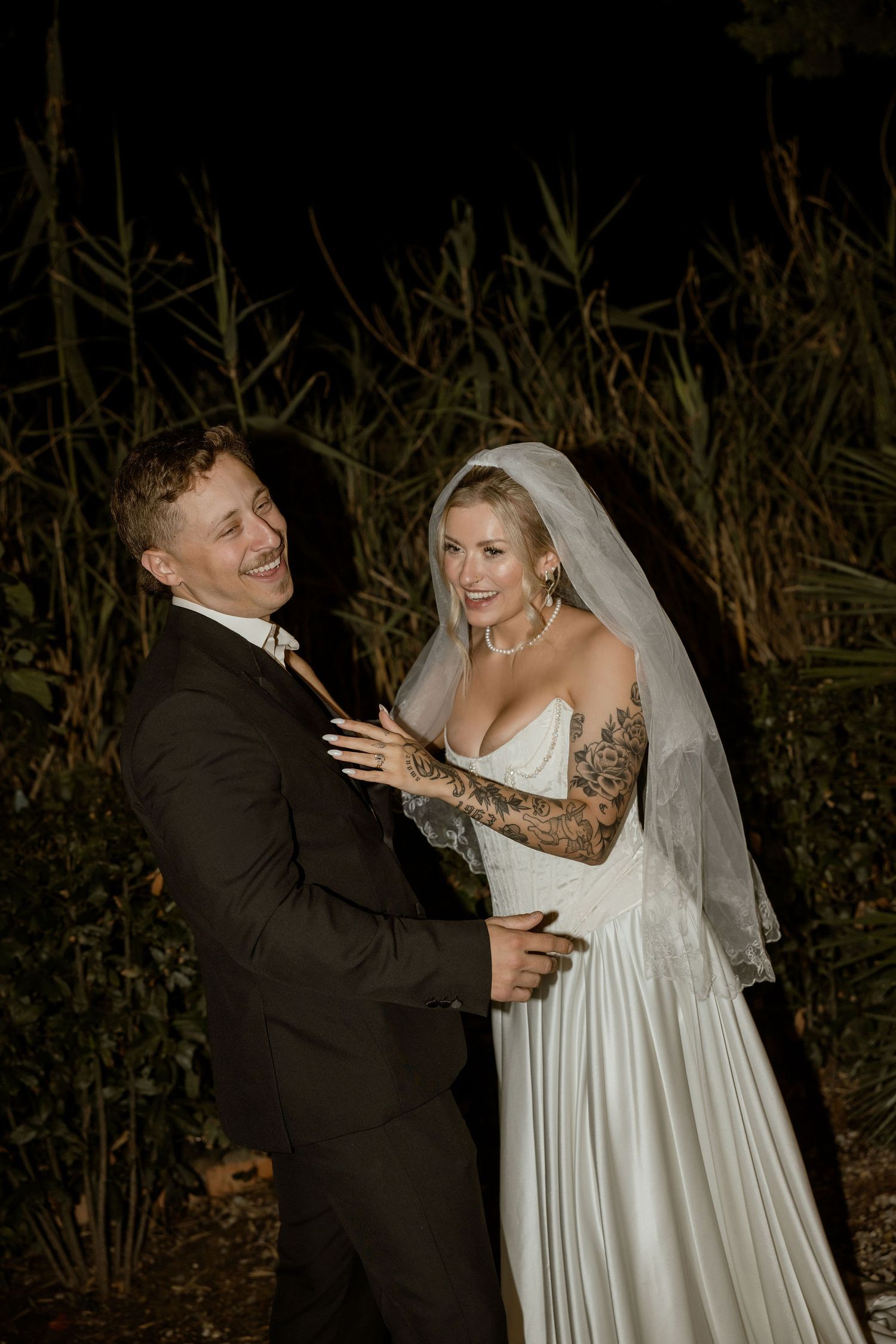 A couple shares a romantic moment in their wedding attire against a dark natural backdrop with tall reeds.