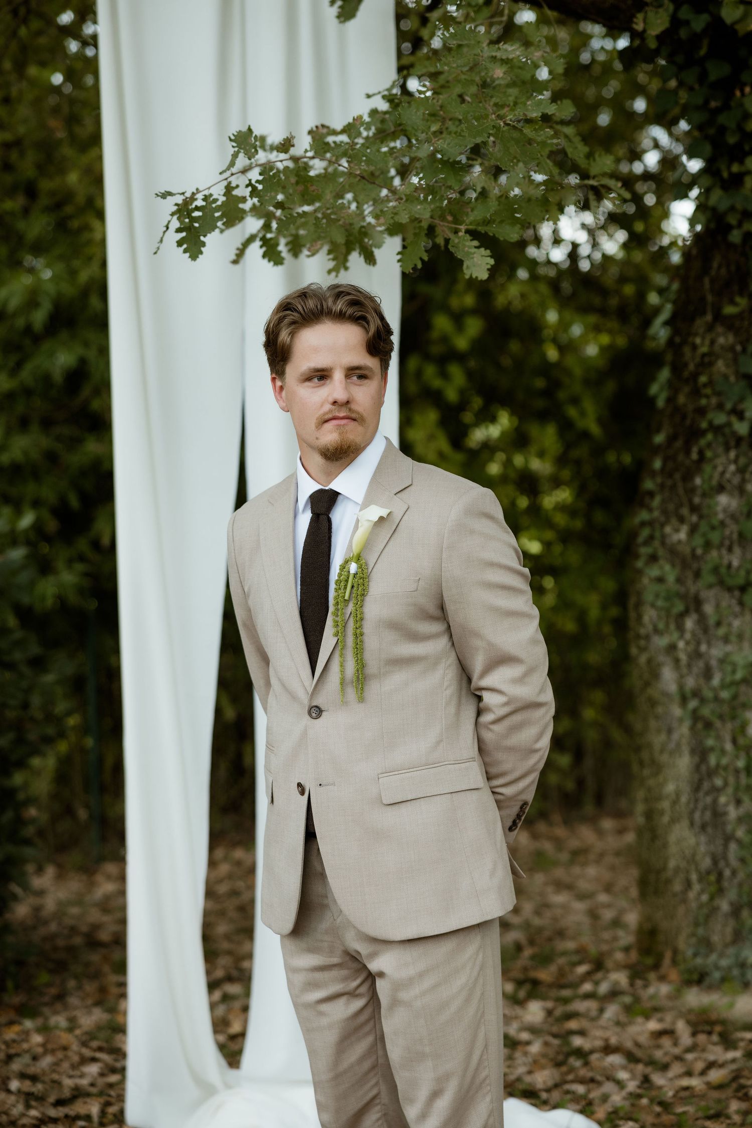 A person in a beige suit stands near white curtains and trees during an outdoor wedding ceremony.