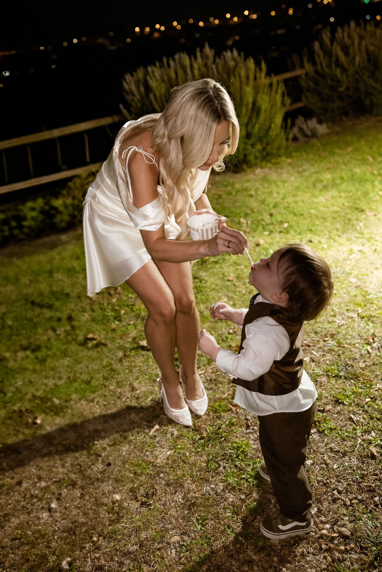 A child in formal wear and woman in white dress share a sweet moment together on grassy ground at night.