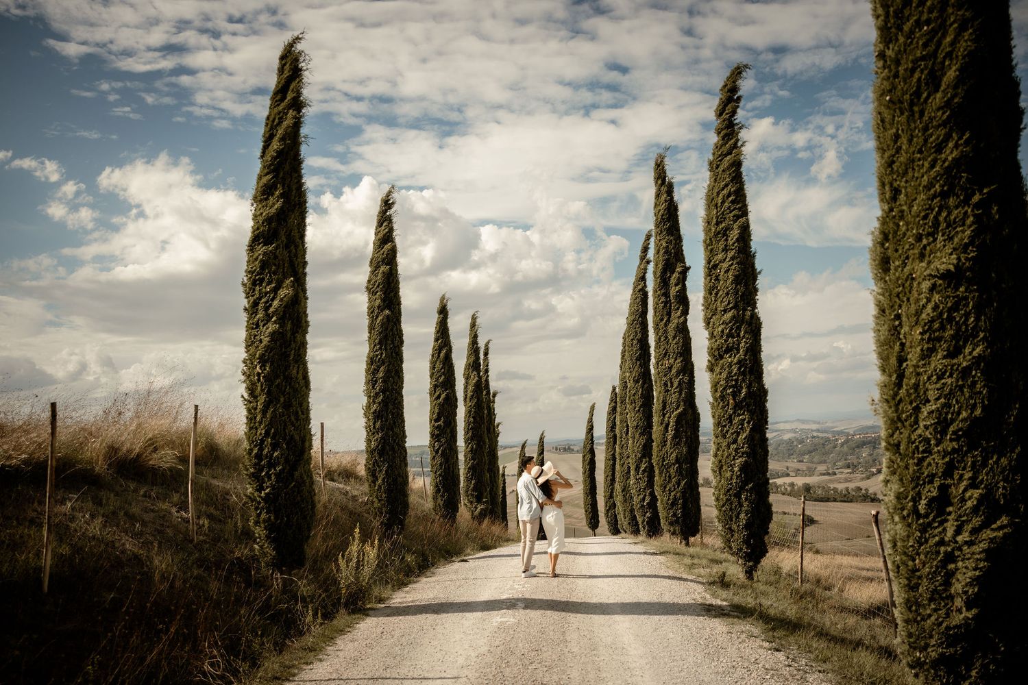 Distant view of couple walking between tall cypress trees under cloudy sky.