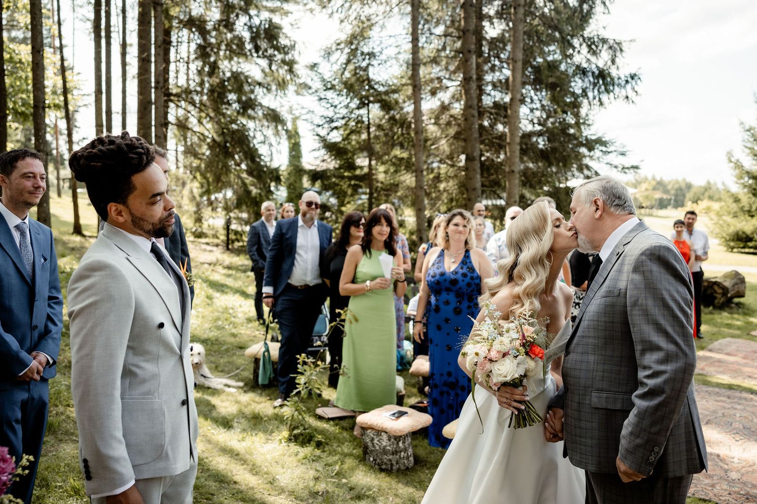 Wedding ceremony in progress among tall trees with guests in formal attire.