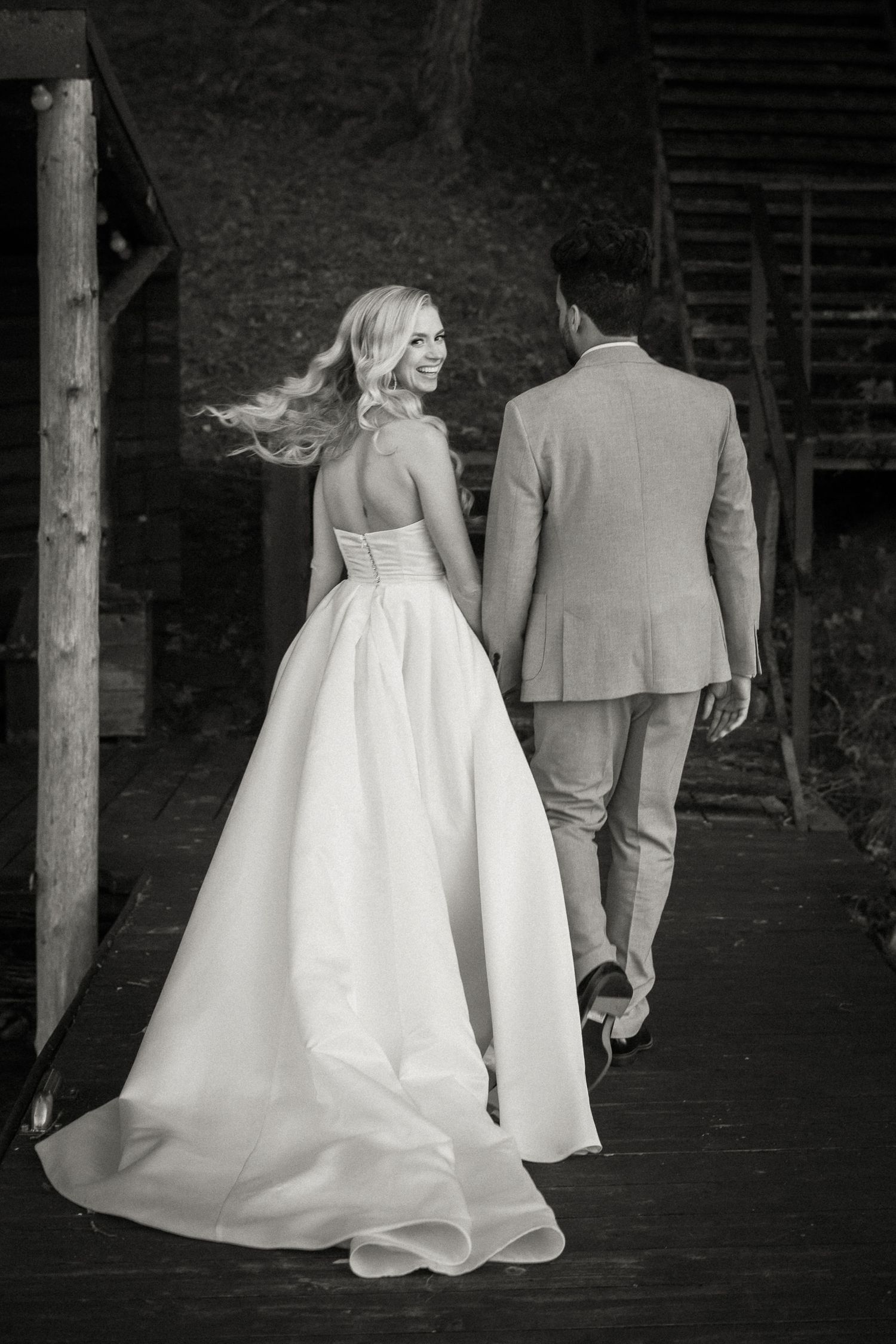 Black and white wedding photograph showing a bride in flowing gown and groom in suit walking on wooden path outdoors.