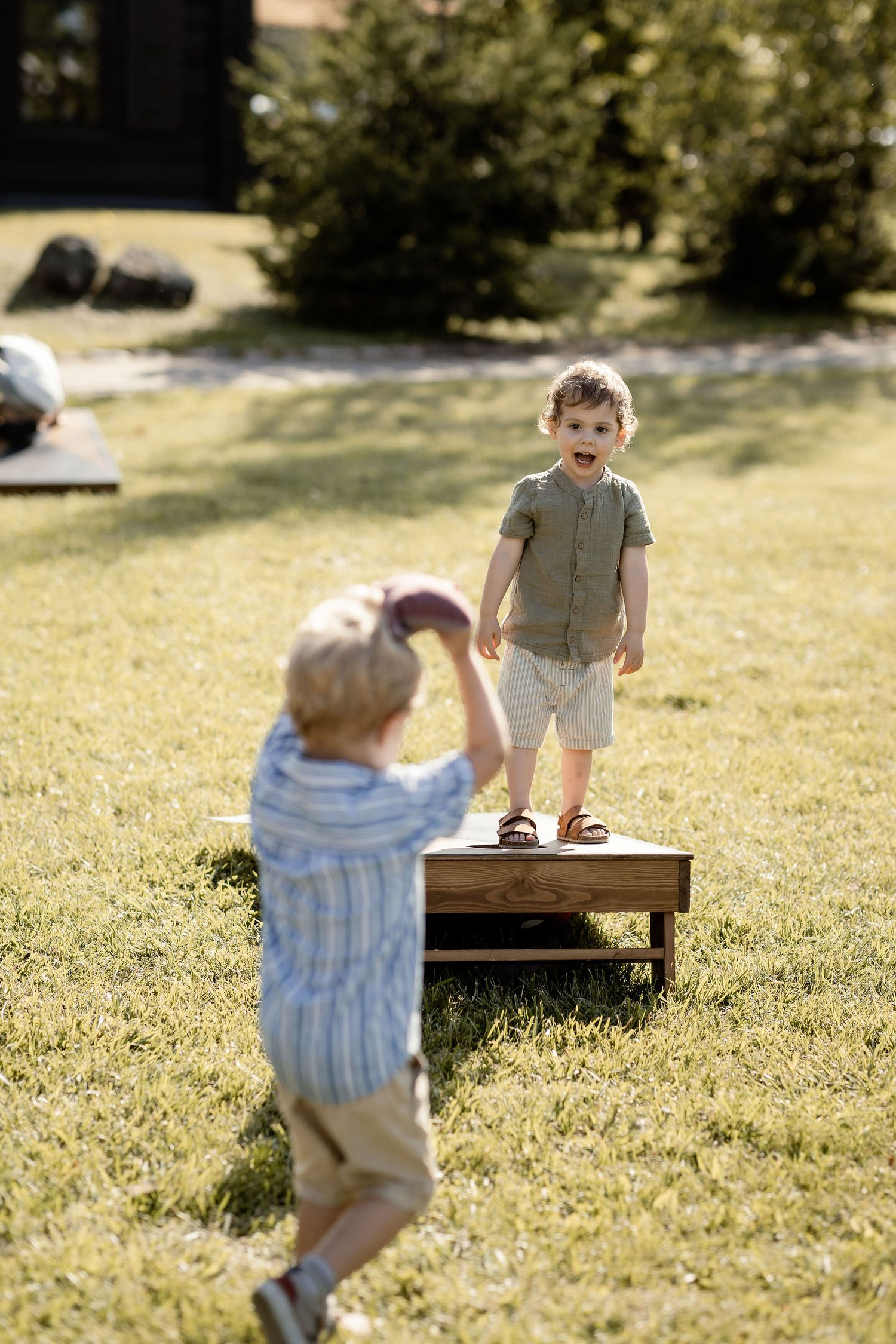 Two children playing outdoors on a sunny day with one standing on a wooden platform.
