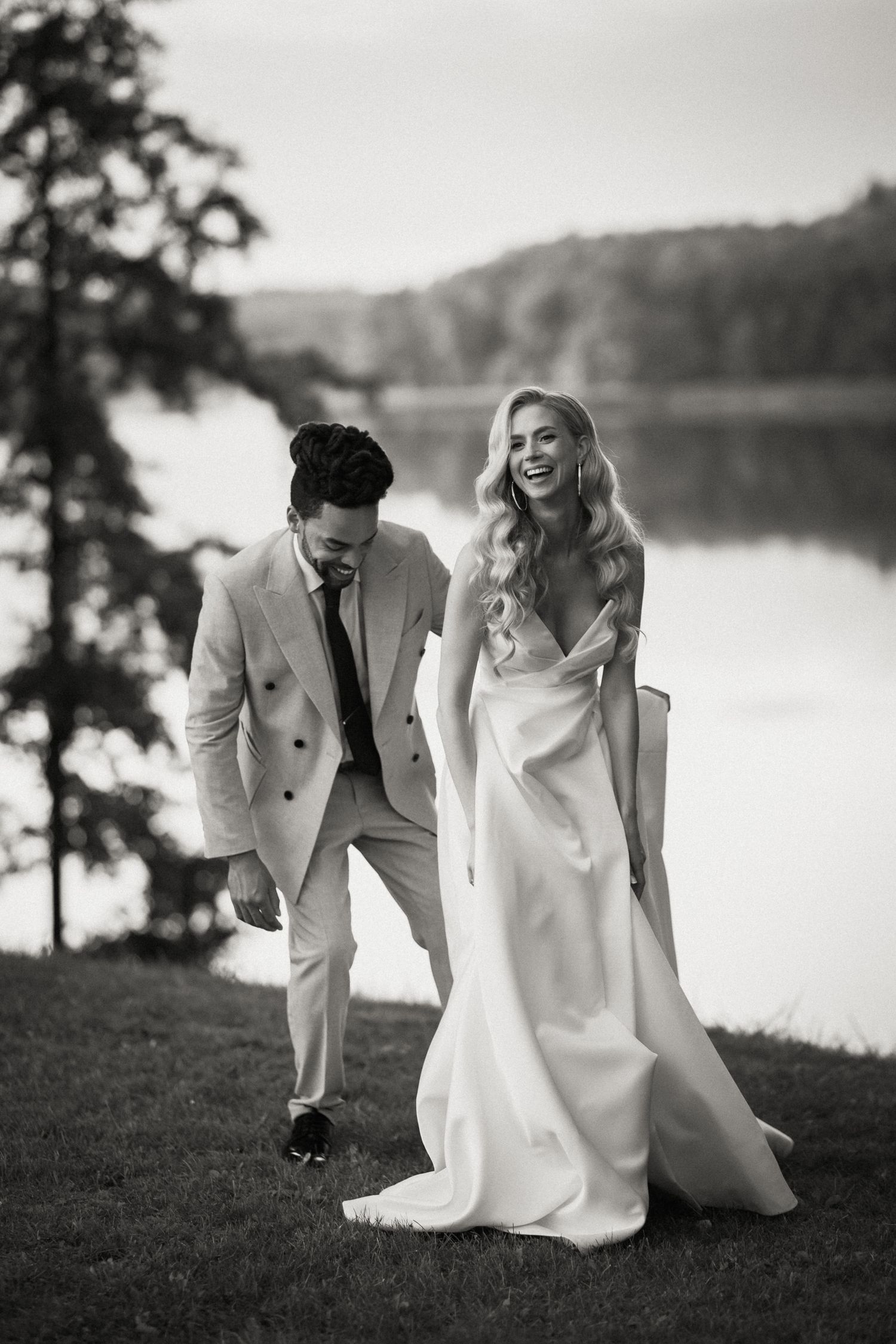 A joyful wedding couple laughs together on a grassy hillside overlooking a serene lake in black and white photography.