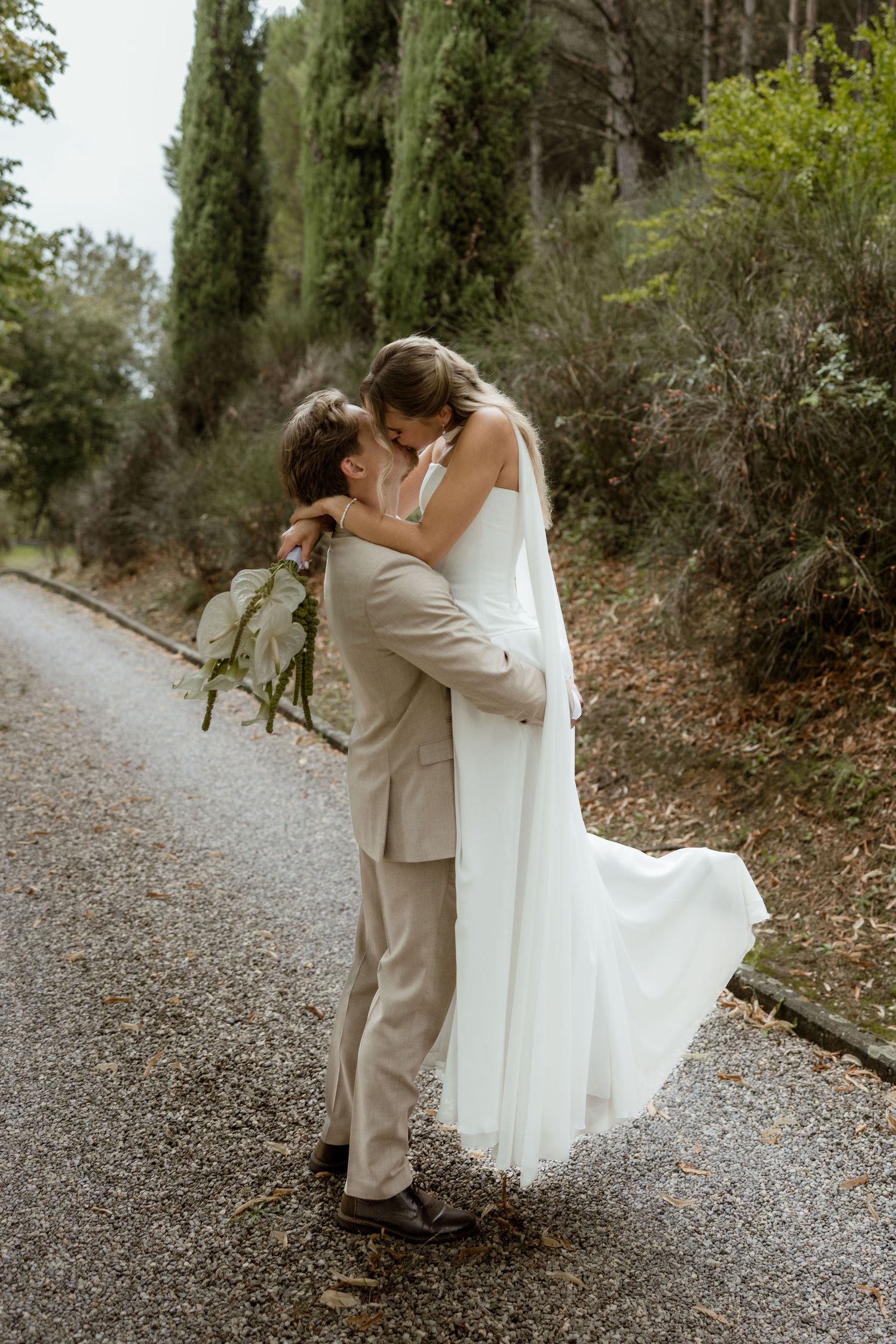 A romantic wedding couple shares an intimate moment on a gravel path surrounded by lush greenery.