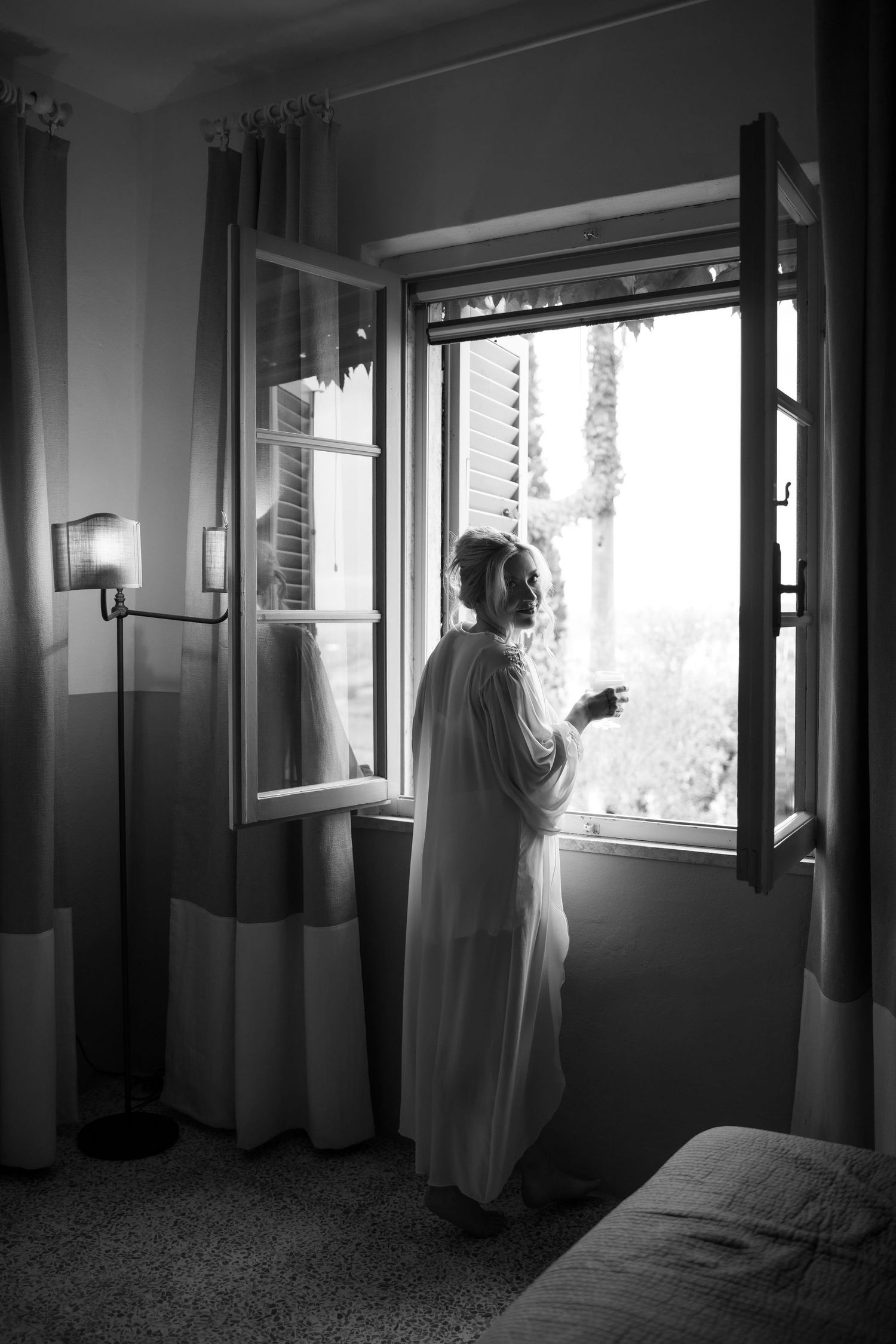A silhouetted figure in a white robe stands at an open window in a moody black and white photograph.