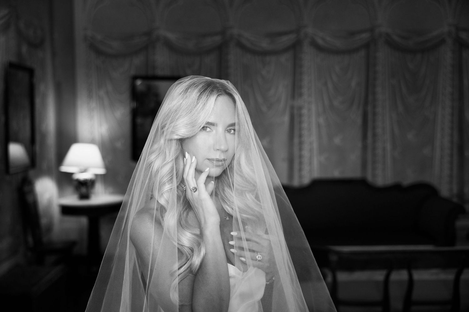 Black and white photo showing a bride in a wedding veil looking down in an elegant indoor setting.