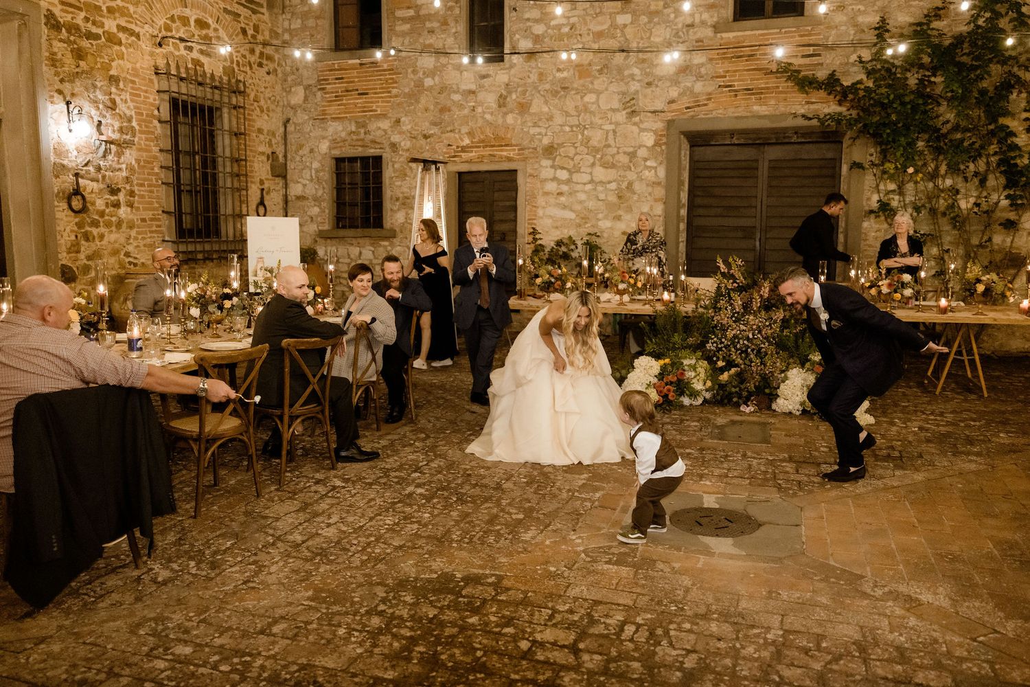 A wedding reception celebration in a rustic stone courtyard with tables and dancing under warm evening lighting.