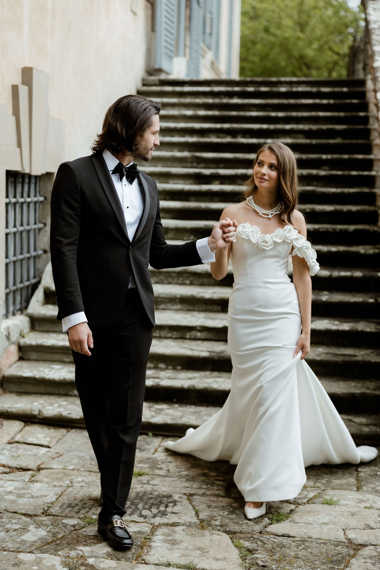 Elegant couple in formal wedding attire descend stone steps together with woman wearing off-shoulder mermaid gown.