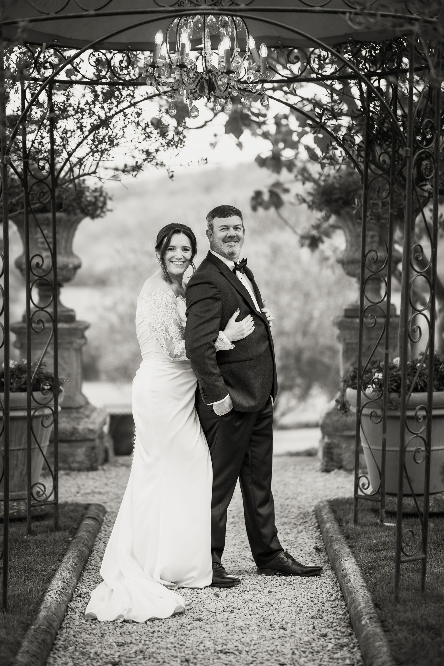 Elegant newlyweds pose under ornate metal archway with chandelier in romantic black and white garden setting.