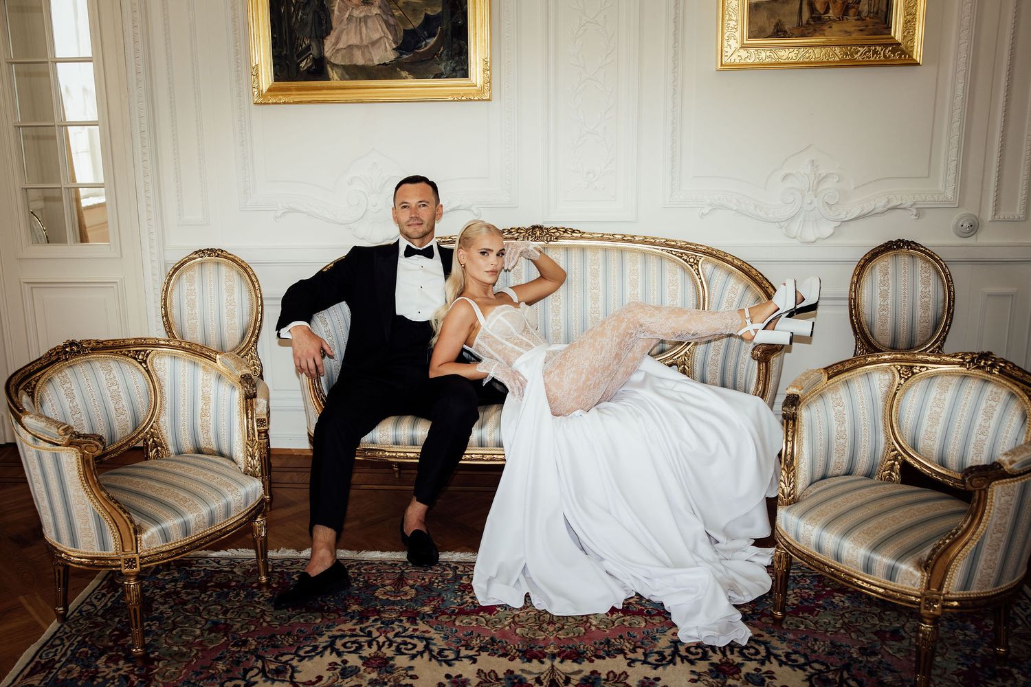 Playful wedding couple relaxes on antique furniture in elegant white room.