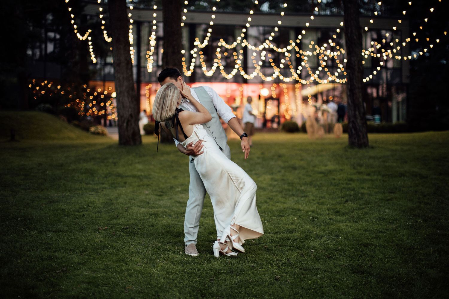A couple dips romantically on a lawn at twilight with string lights and trees creating a magical outdoor setting.