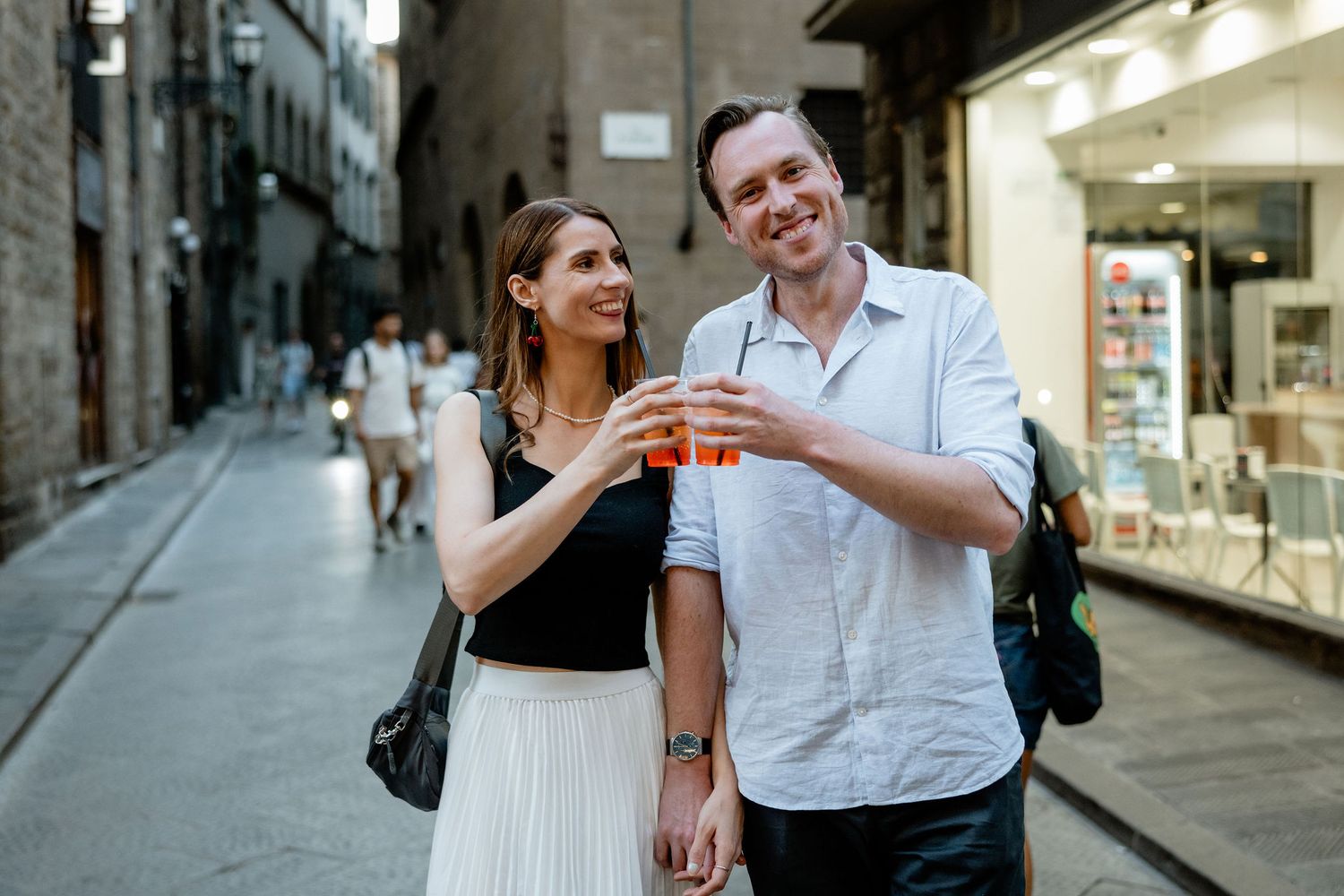 A couple enjoys drinks while walking down a narrow cobblestone street in Italy with storefronts in the background.