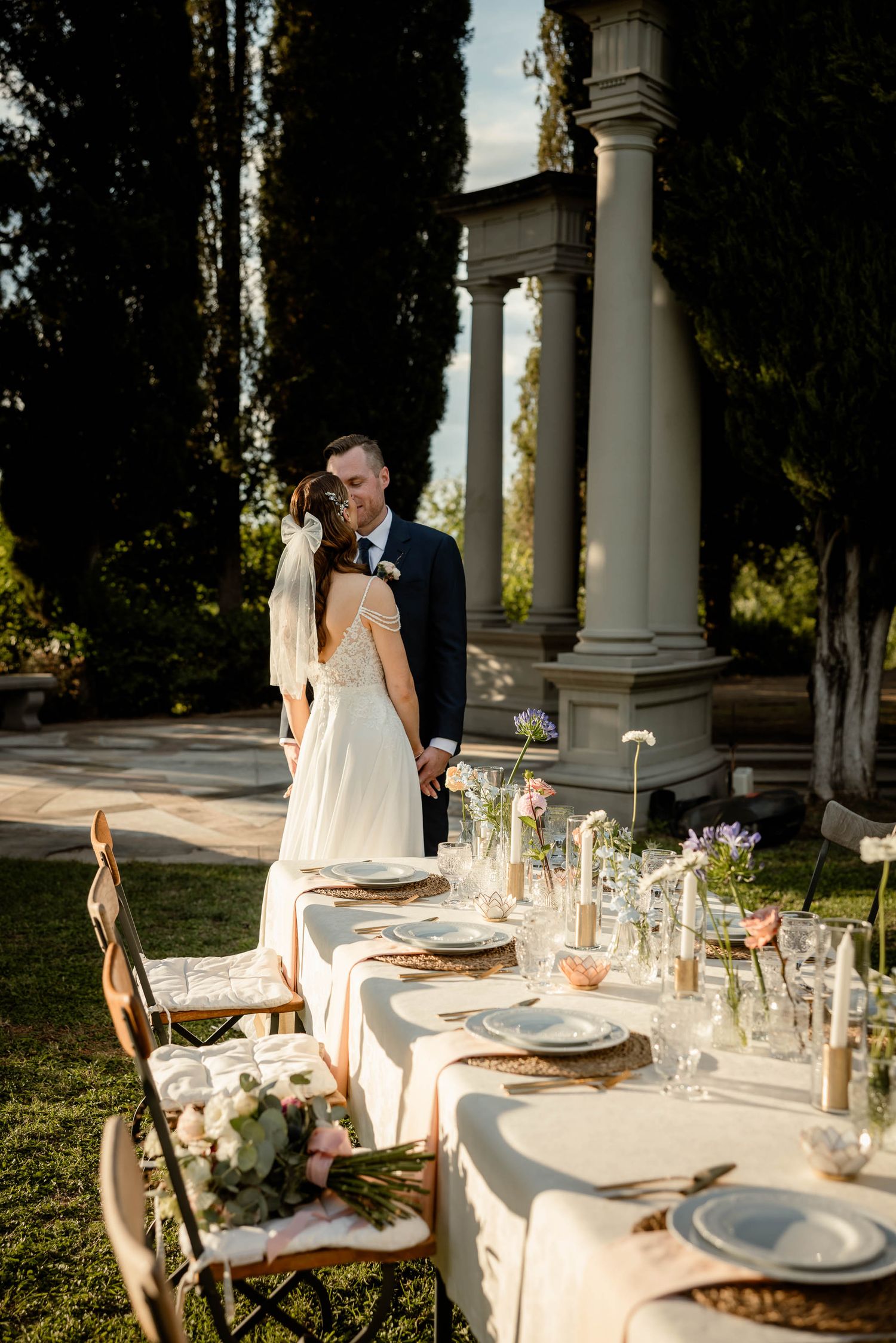 Elegant wedding table setting with white linens and place settings beneath Roman columns in an outdoor garden.