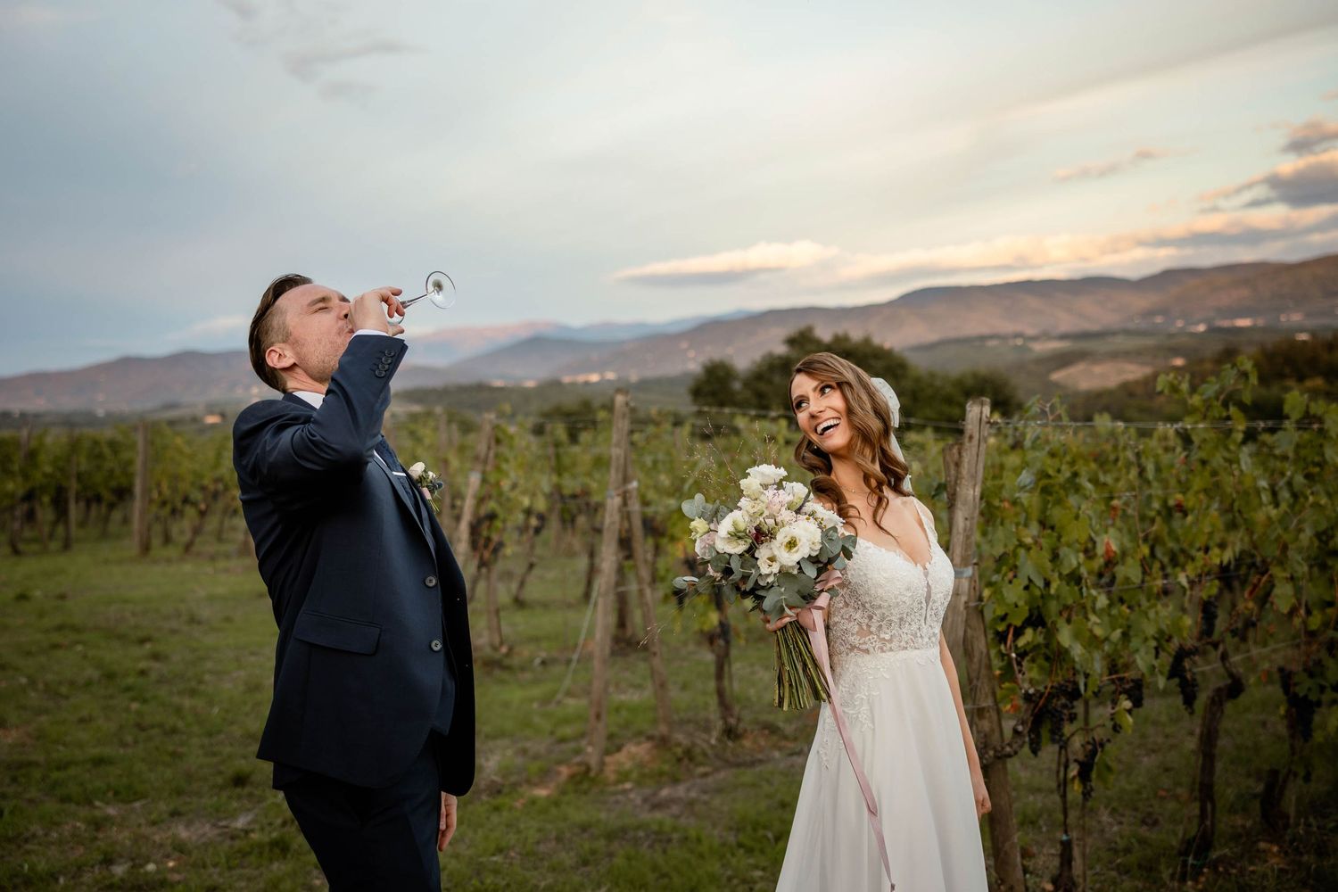 Wedding couple poses playfully in a vineyard during golden hour with mountains in the background.