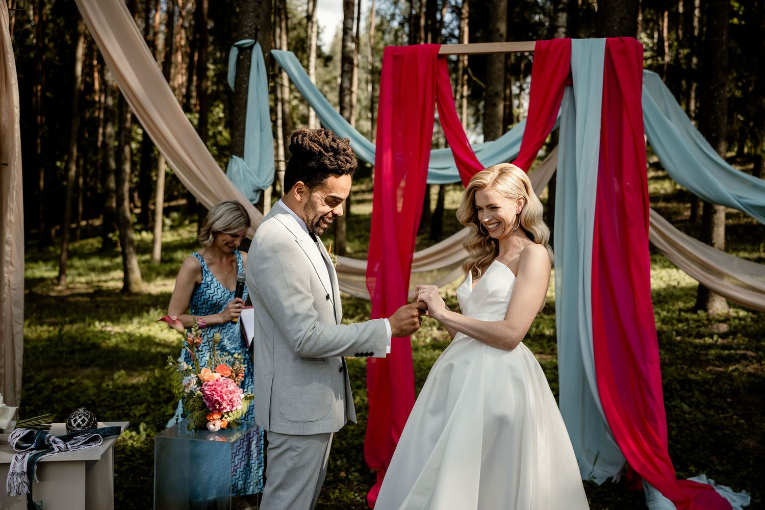 Wedding ceremony in forest with red and blue fabric draping creates romantic outdoor setting for exchanging rings.