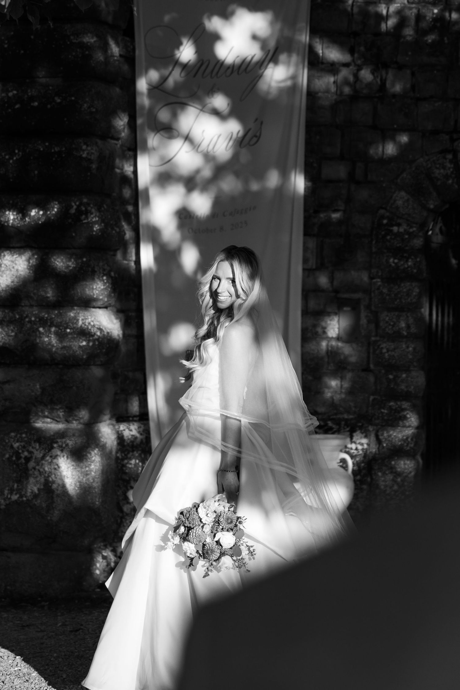 A white wedding dress hangs elegantly in dappled sunlight creating dramatic shadows and highlights against a dark wall.
