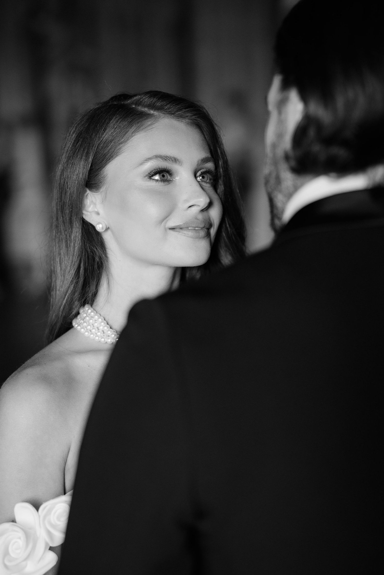 A bride in a pearl choker necklace smiles while looking over her shoulder in an elegant black and white portrait.