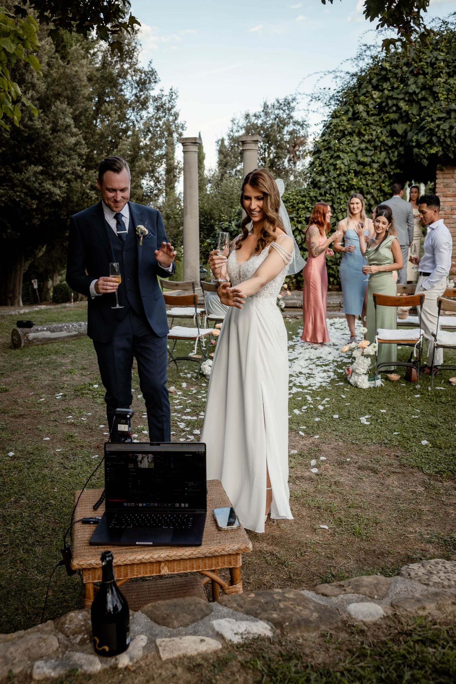 Couple toasting with champagne at outdoor wedding with vintage columns and gathered guests.