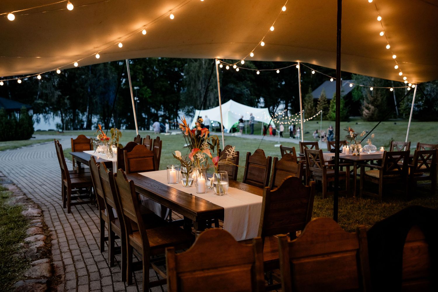 Rustic outdoor wedding reception under a tent with string lights and wooden chairs on a stone patio at dusk.