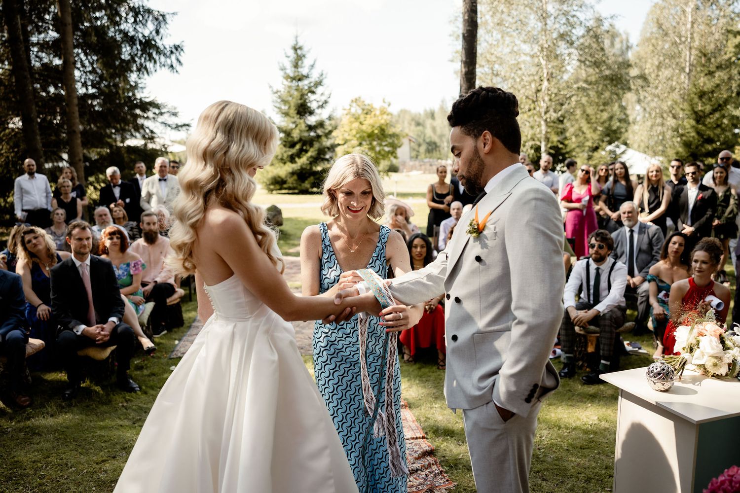 An outdoor wedding ceremony takes place in a forest clearing with guests watching.